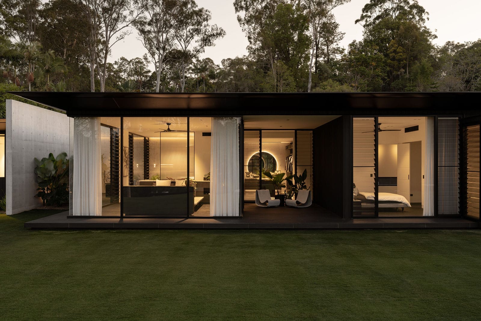 Blackwood by Sarah Waller Architecture. Photography by Alyne Media. Pavilion home at night with warm light emanating. Grass in foreground. Floor-to-length windows with full-length white curtains. 