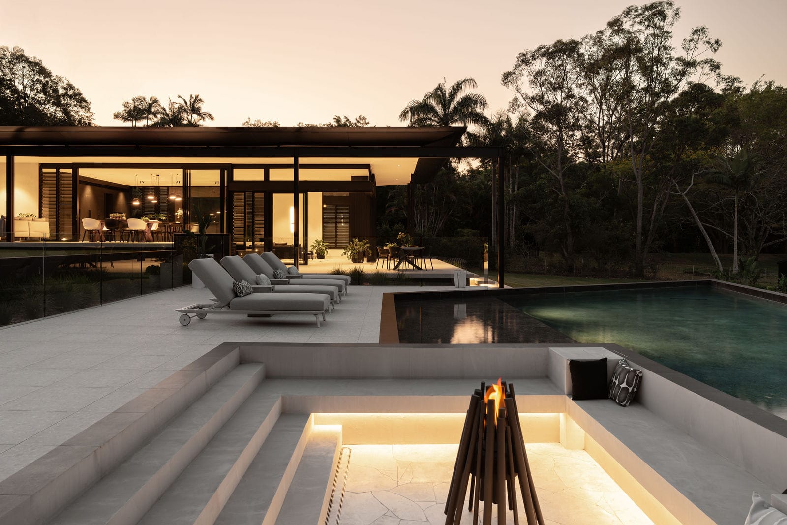 Blackwood by Sarah Waller Architecture. Photography by Alyne Media. Outdoor patio area with raised concrete pool and day beds. Sunken firepit in foreground, home and tropical trees in background. 