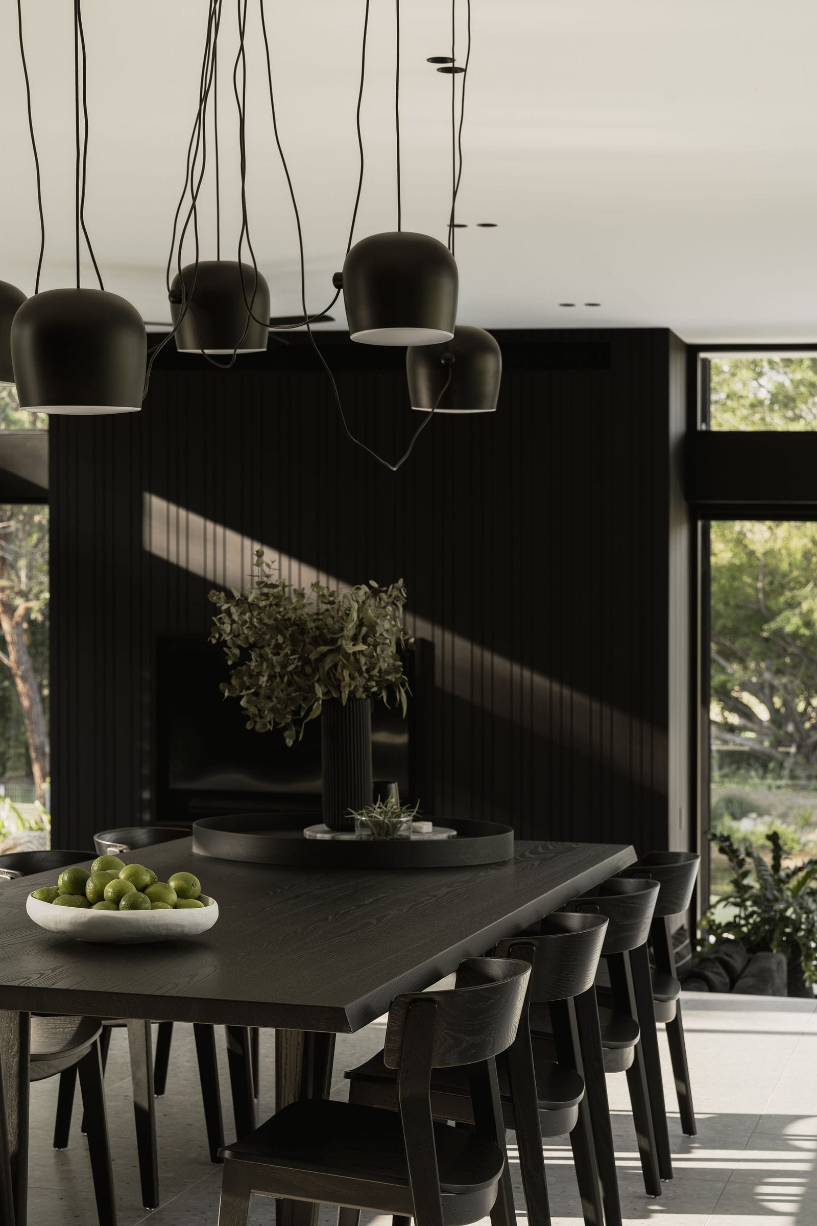 Blackwood by Sarah Waller Architecture. Photography by Alyne Media. Black timber dining table and chairs with multiple black pendant lights hanging overhead. Black timber clad in background with windows on either side. 
