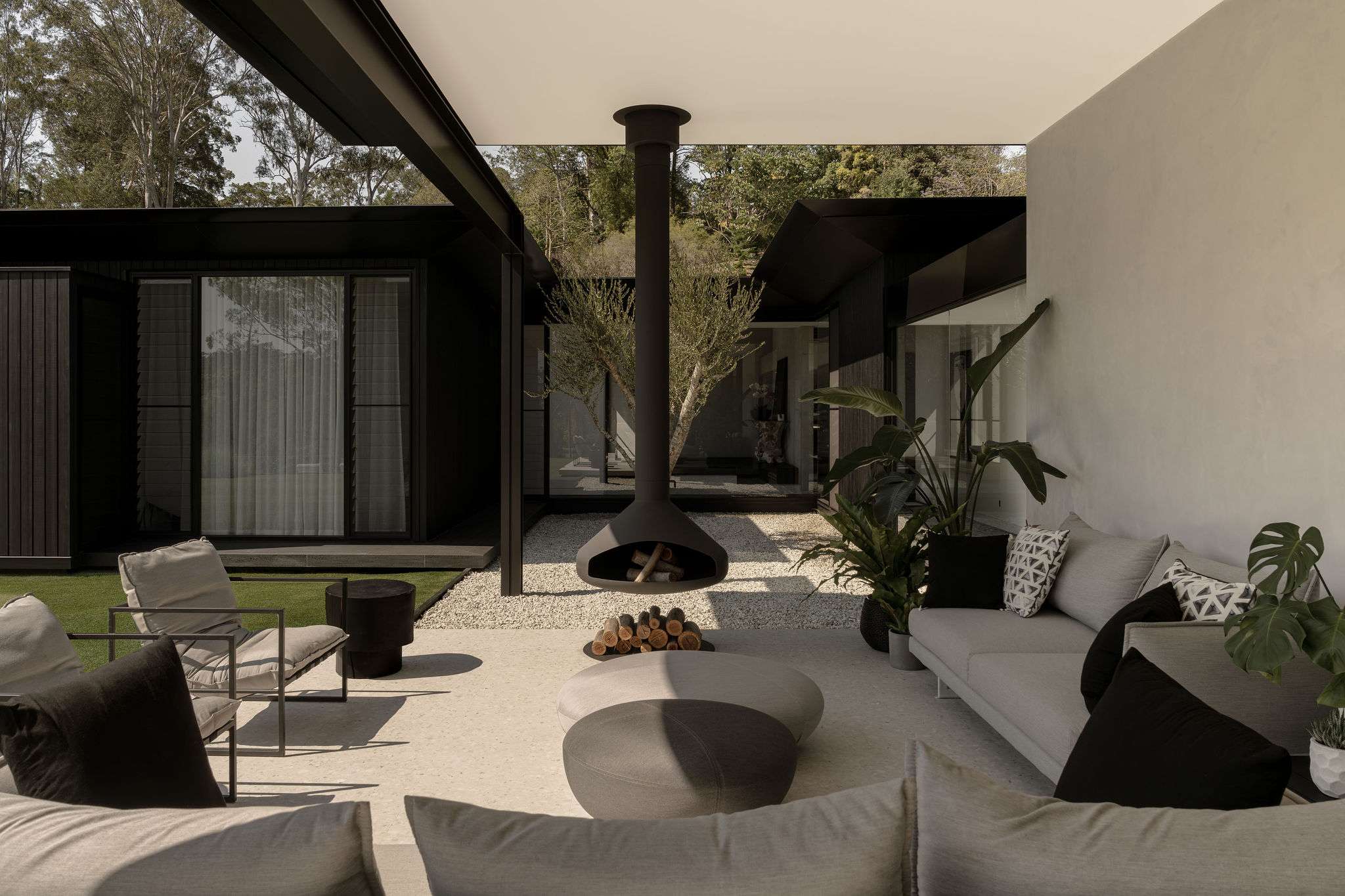 Blackwood by Sarah Waller Architecture. Photography by Alyne Media. Outdoor courtyard with roof mounted black fireplace surrounded by grey couches and armchairs and soft coffee tables. 
