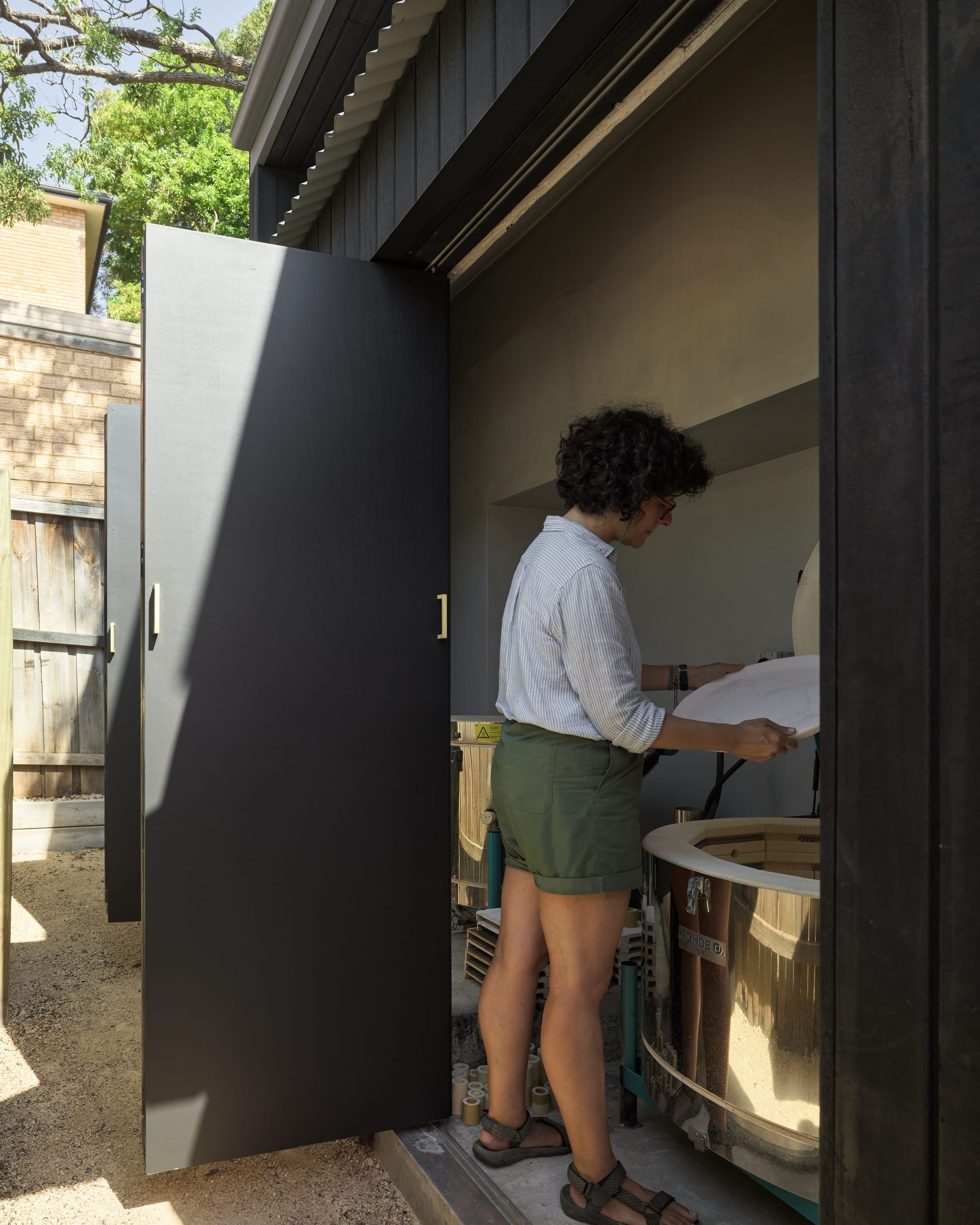 Coconut Crab by Alexander Symes Architect. Photography by Barton Taylor Photography. Woman standing in front of steel kiln. Dark exterior walls and door.
