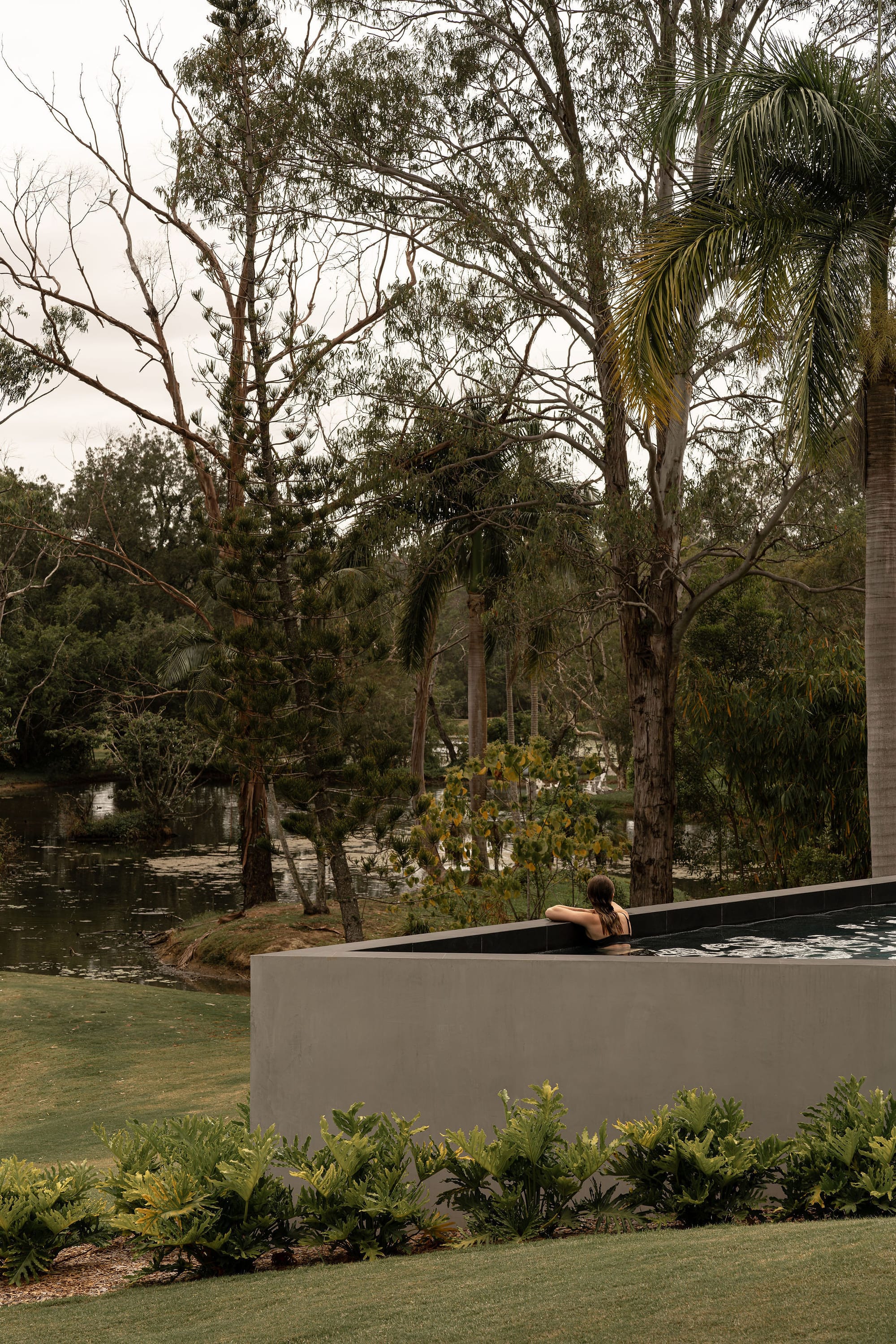 Blackwood by Sarah Waller Architecture. Photography by Alyne Media. Outdoor verandah with concrete floors and black beams and pillars. Pool in background, rolling green hills to left. 