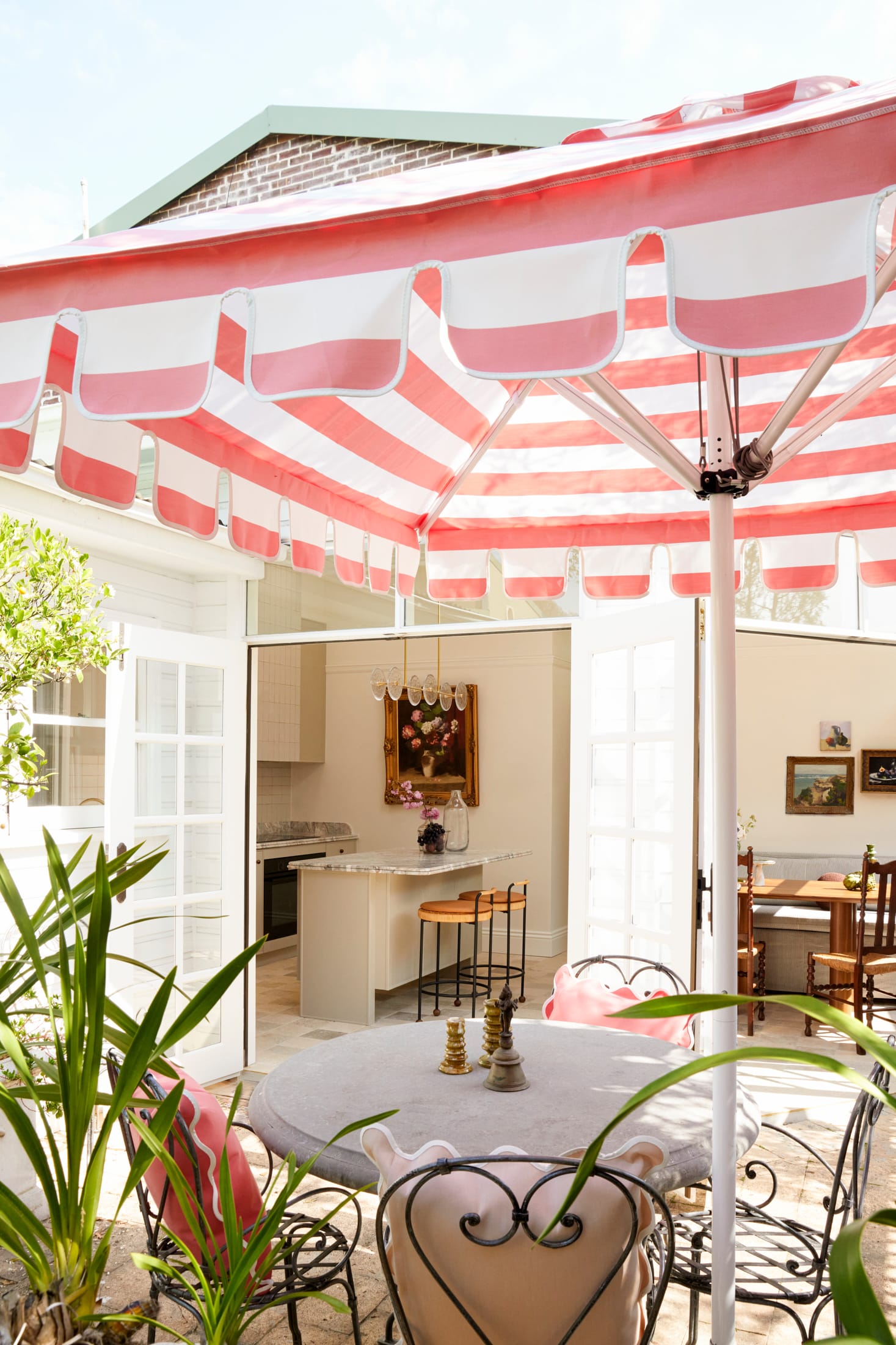 Drummoyne House by studio BARBARA. Photography by Jacqui Turk.  Outdoor dining setting with large pink and white striped umbrella. Wrought iron vintage dining chairs surrounded by green plants.