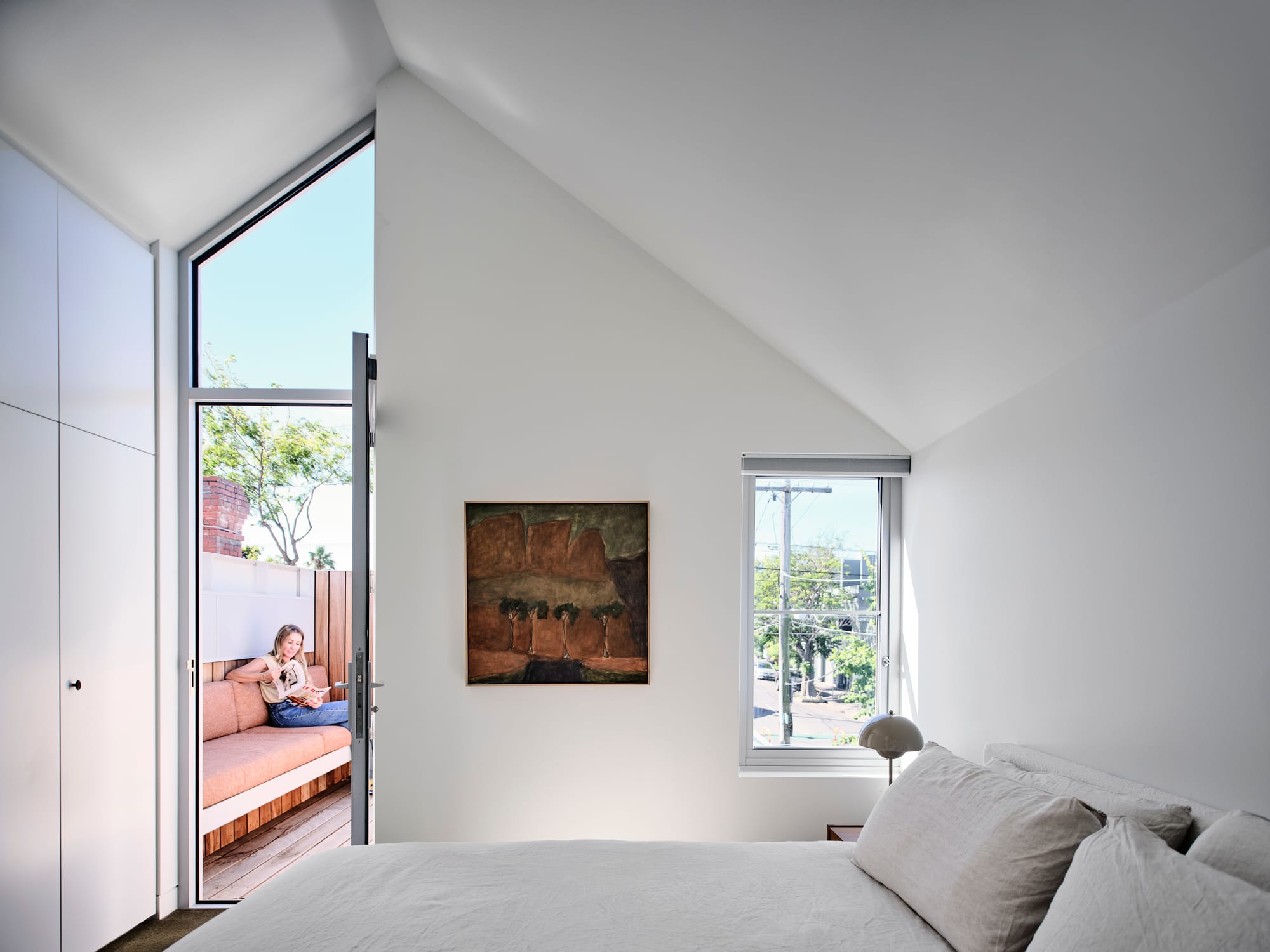 House Northroy by True Story. Photography by Dean Bradley. Bedroom with white pitched ceilings and white bed linen. Door opens onto outdoor terrace with timber bench seat.