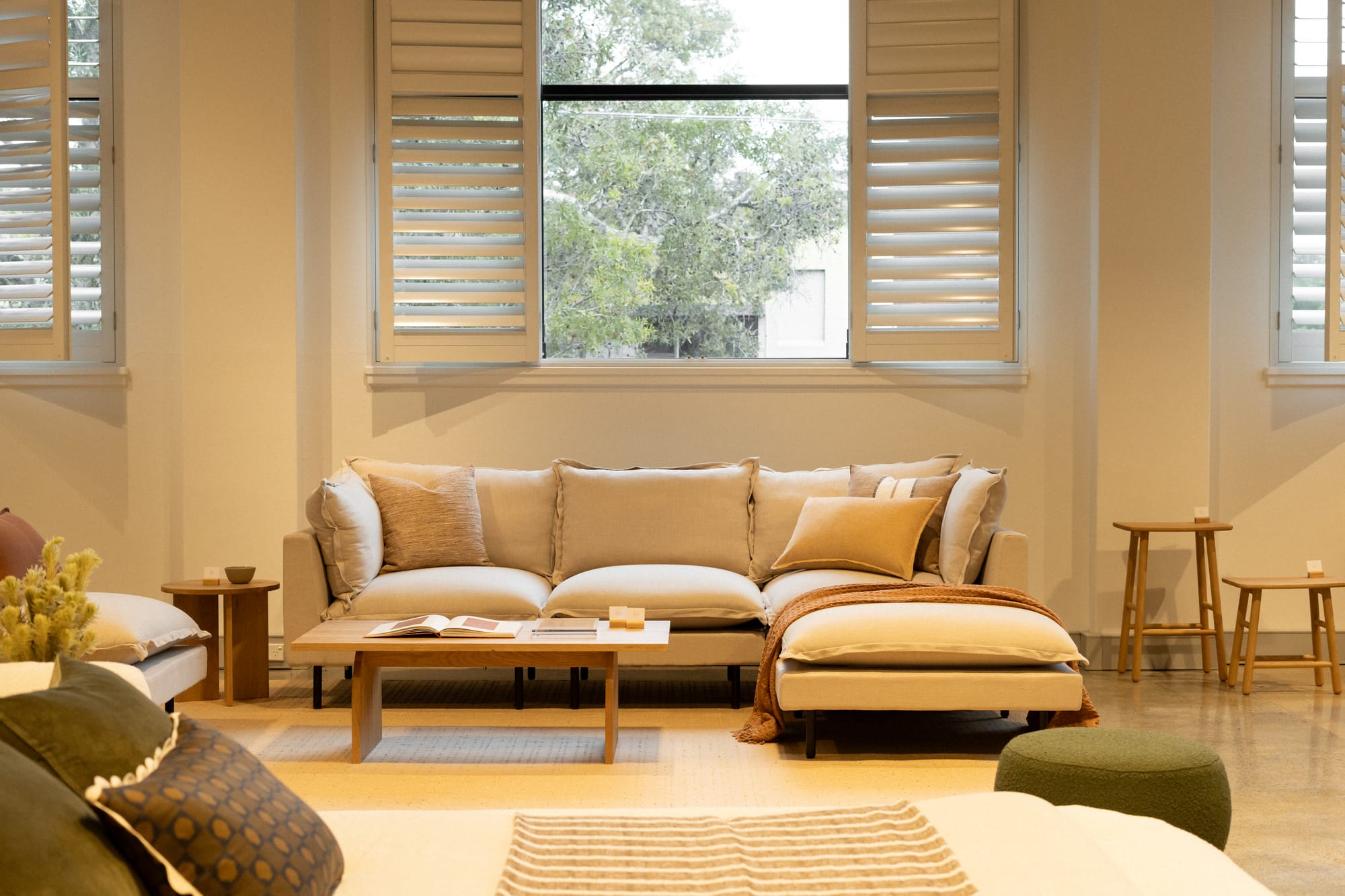 RJ Living's New Sydney Retail Space. Photography by Nathan Oakley. White modular lounge below window with white plantation shutters. Timber coffee table. Bed with green throw cushions in foreground.
