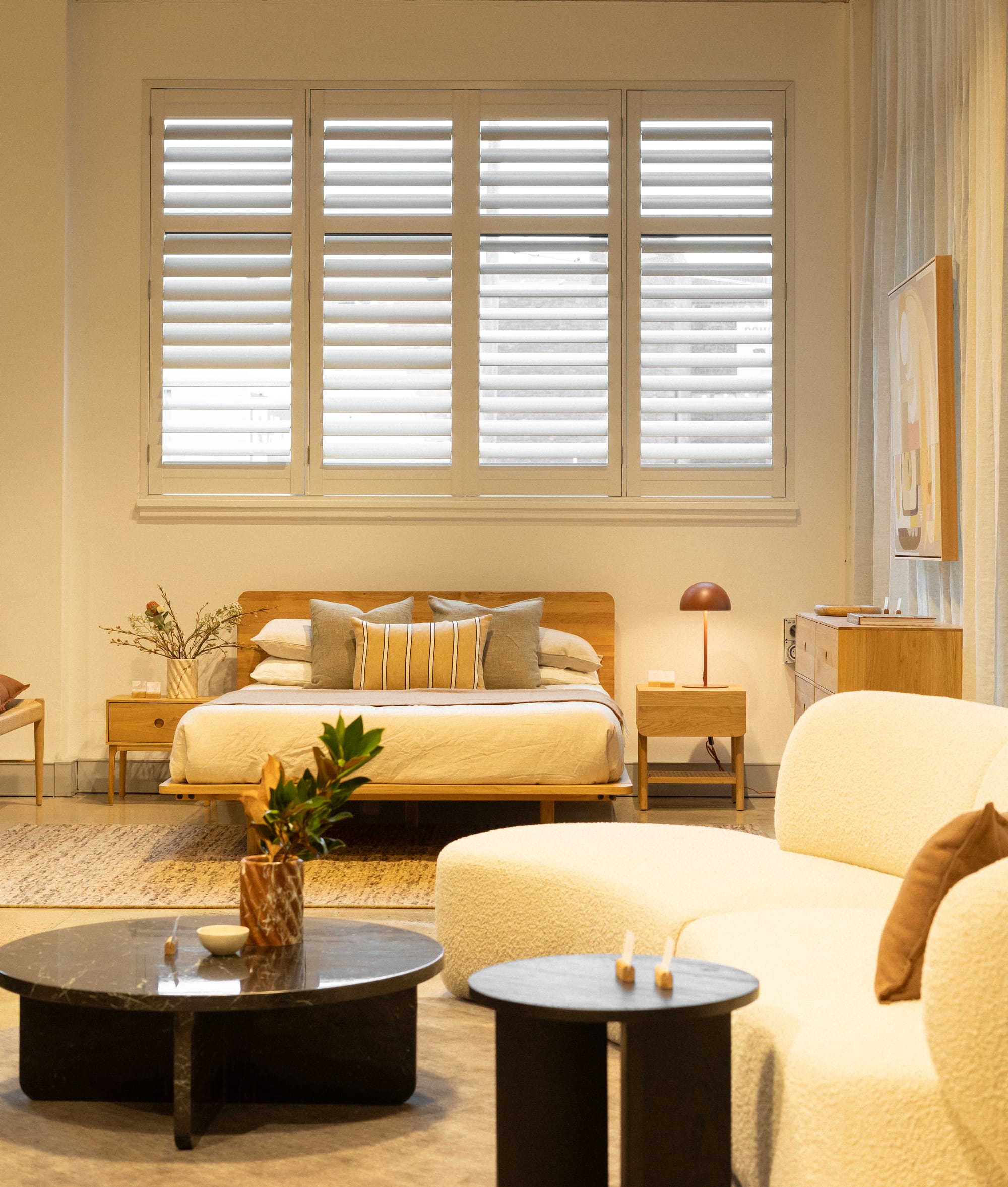 RJ Living's New Sydney Retail Space. Photography by Nathan Oakley. Showroom space with simulated bedroom and living spaces. Timber framed bed with white linen and matching bedside tables in background. White boucle couch with black stone coffee table in foreground. Large window with plantation shutters above bed.