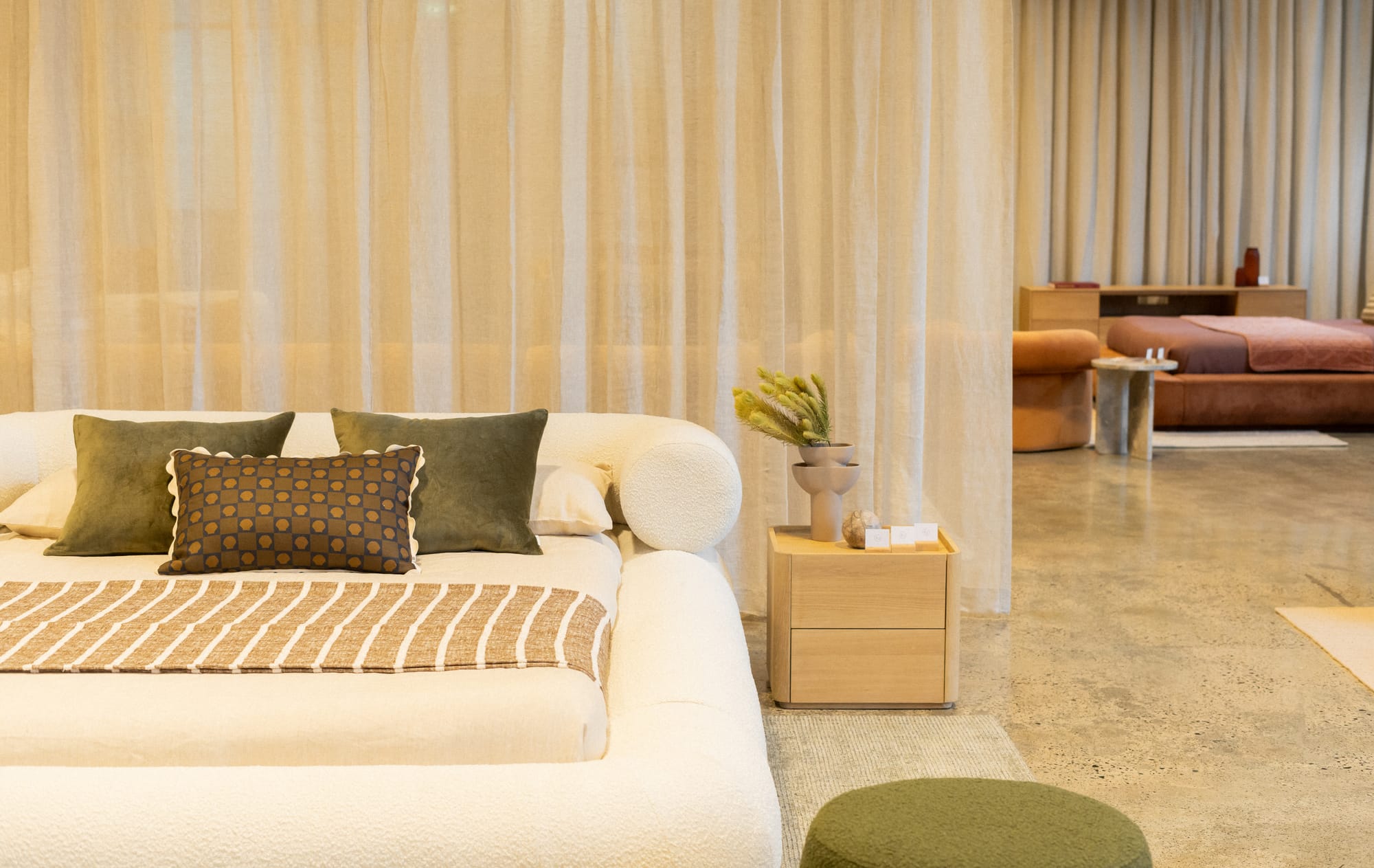 RJ Living's New Sydney Retail Space. Photography by Nathan Oakley. White boucle bed frame with green throw cushions. Polished concrete floors and floor-to-ceiling sheer curtains. Timber bedside table and green floor pouf.