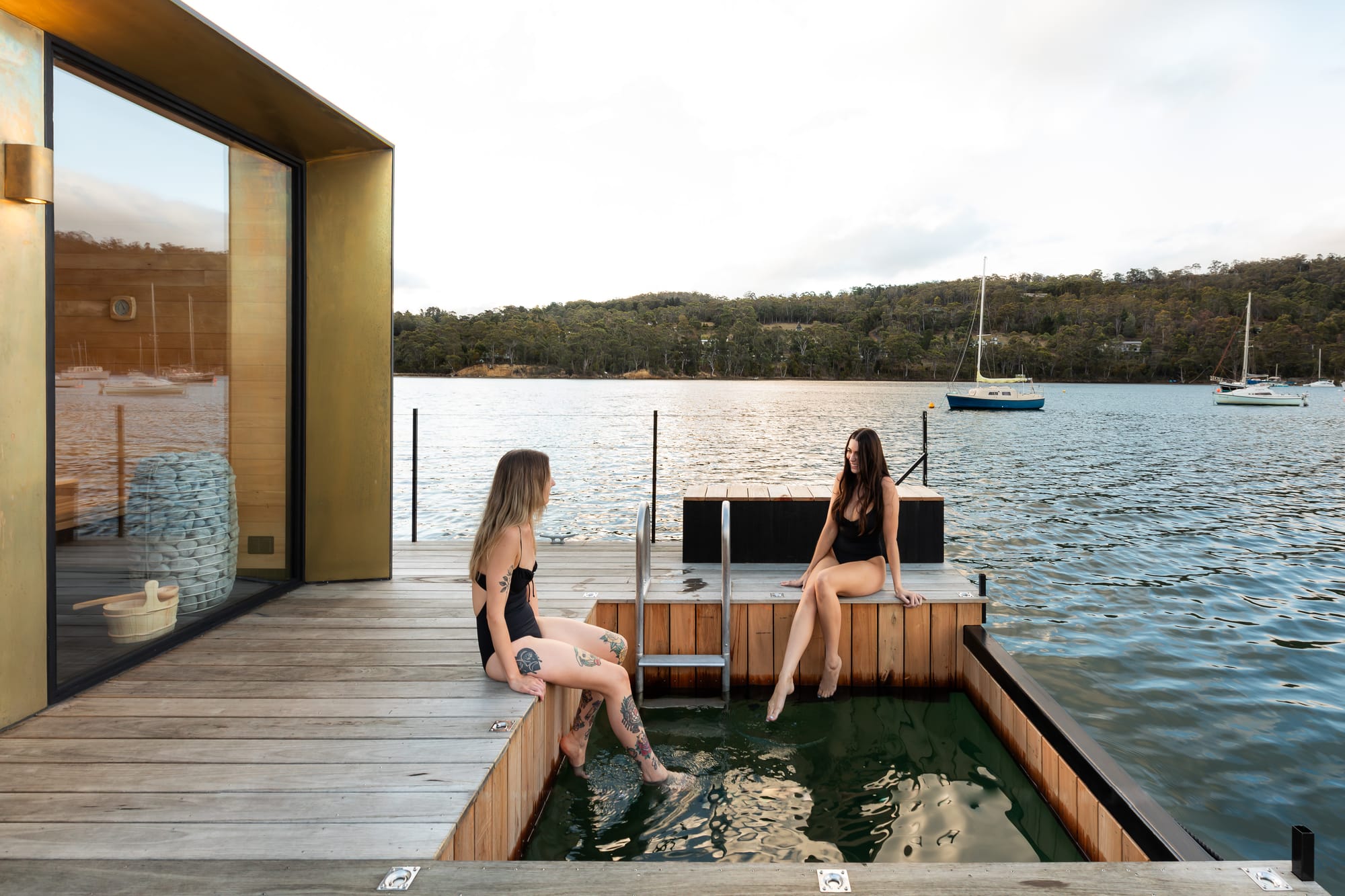 Sauna Boat Tasmania. Photography by Natasha Mulhall. Two women with feet in outdoor freshwater pool in outdoor timber deck. Surrounded by large body of water. Bronze metal sauna to left. 