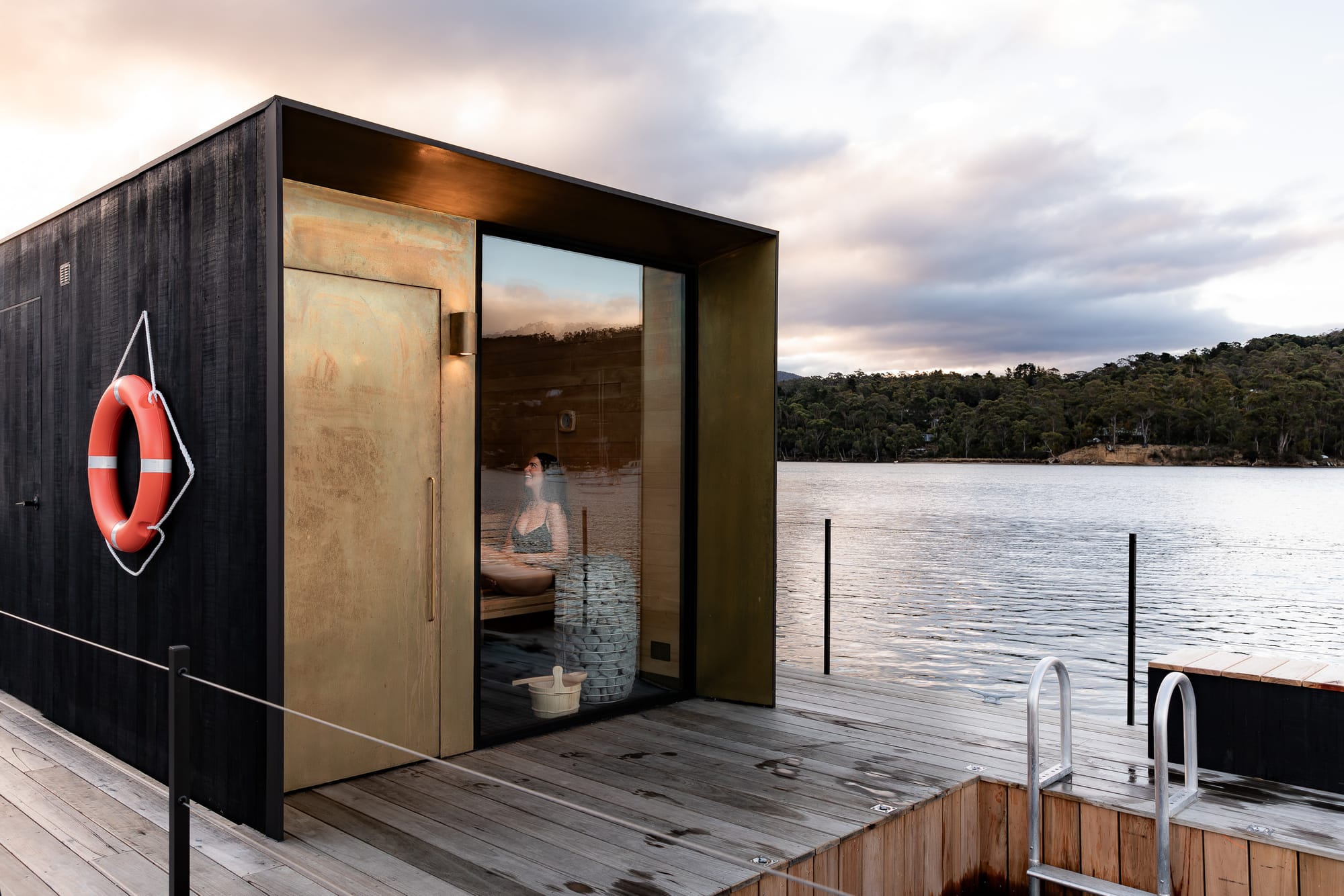 Sauna Boat Tasmania. Photography by Natasha Mulhall. Black timber and bronze sauna boat with timber deck and outdoor pool. Floating on large body of water, hills in background. 