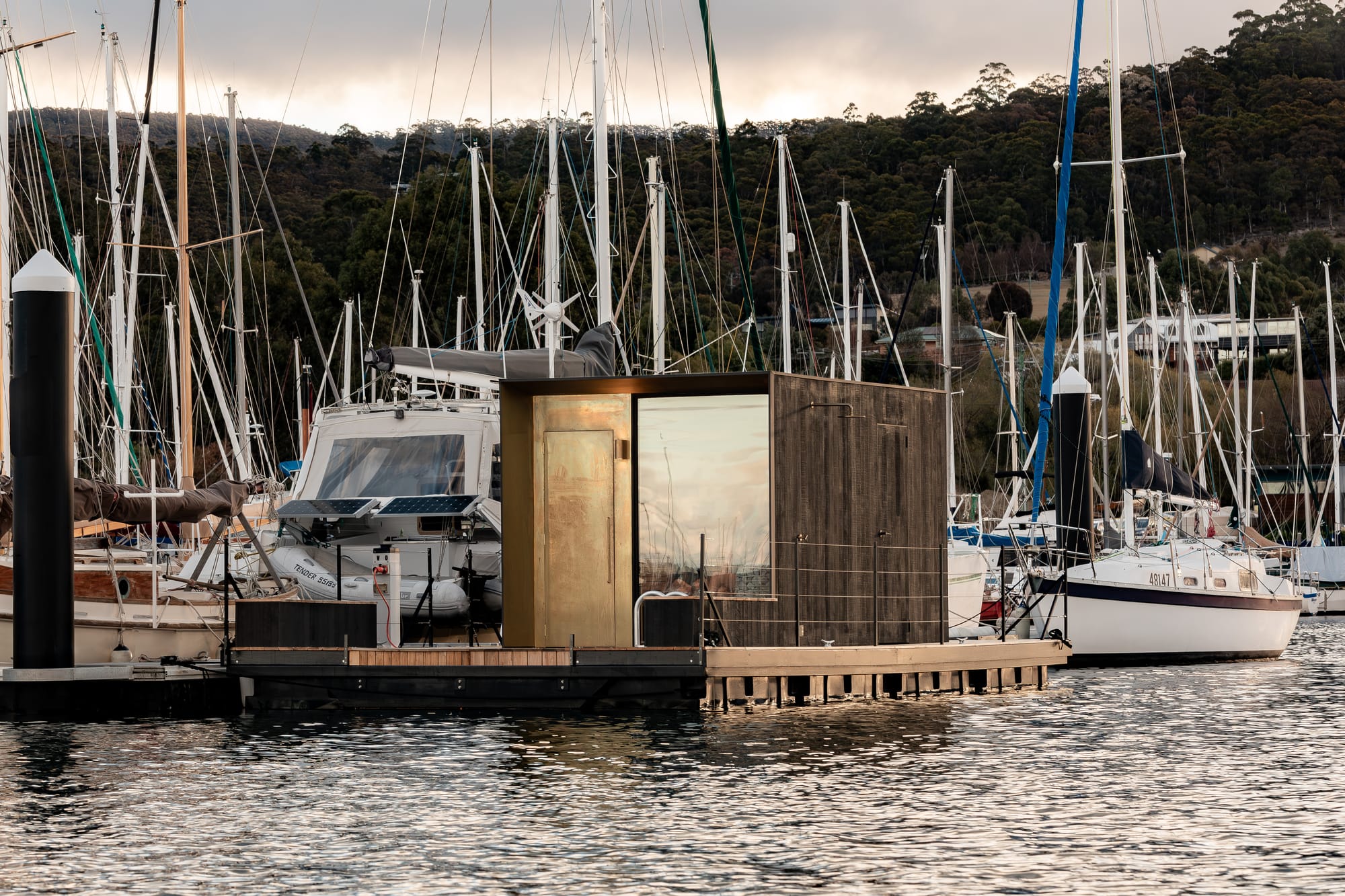 Sauna Boat Tasmania. Photography by Natasha Mulhall. Sauna boat in harbour surrounded by lots of other boats. Hills in background. 