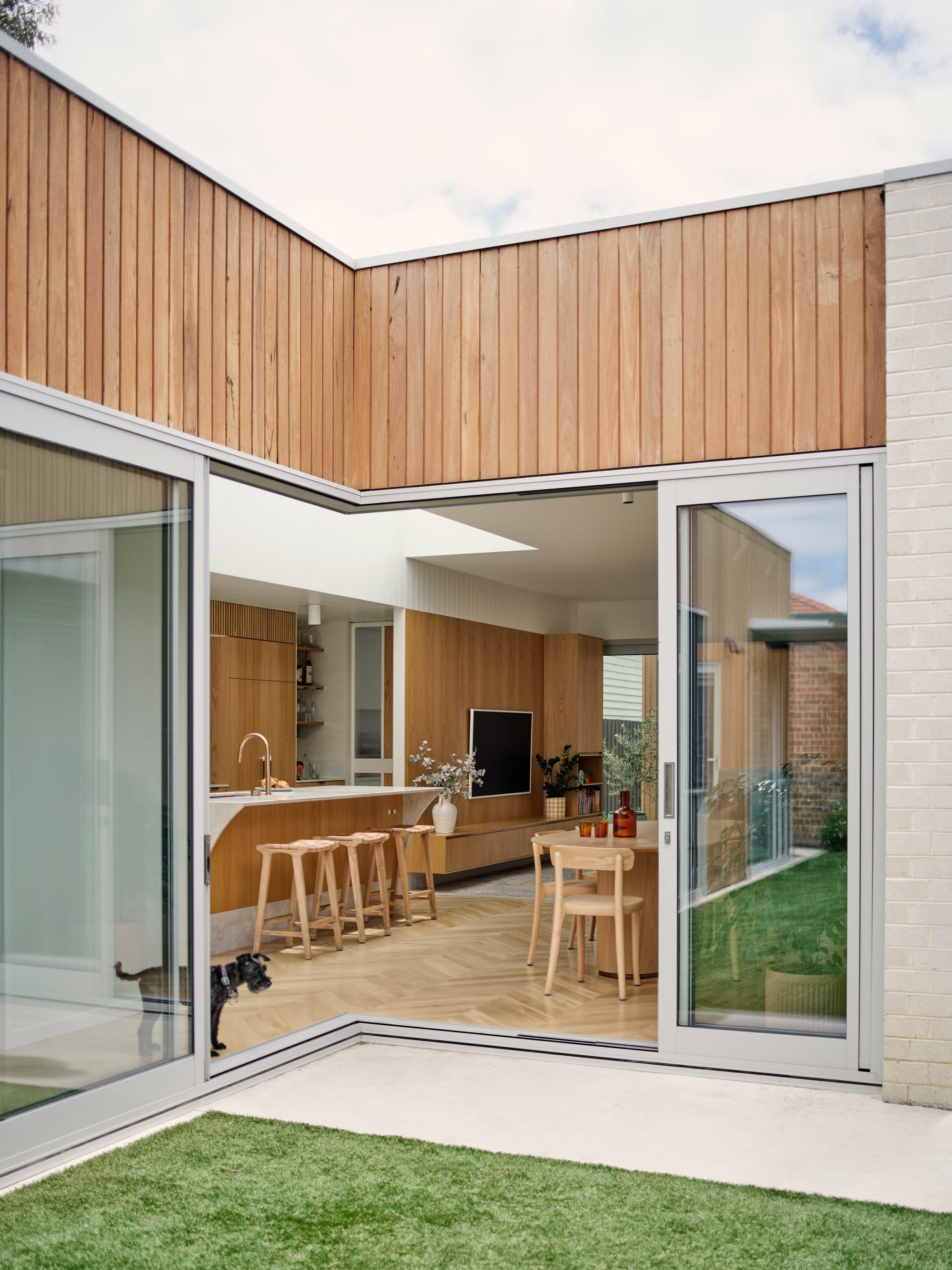 ZigZag House by True Story. Photography by Dean Bradley. Living, dining and kitchen with timber veneer walls and cabinetry, herringbone floors and furniture. Opens onto grass courtyard.