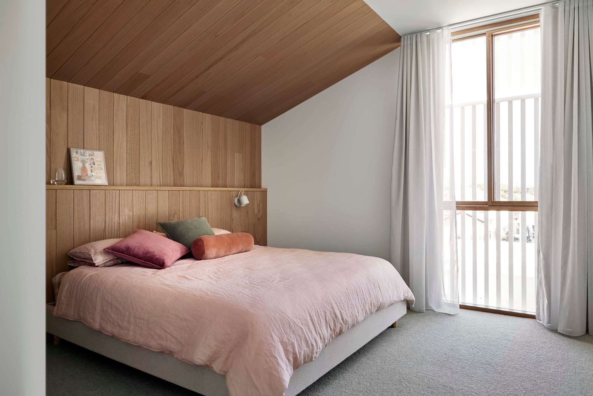 Rosie by Eckersley Architects. Photography by Tess Kelly. Timber walls and ceiling in bedroom with grey carpets and floor to ceiling sheer curtains. Pink bed linen and white walls. 