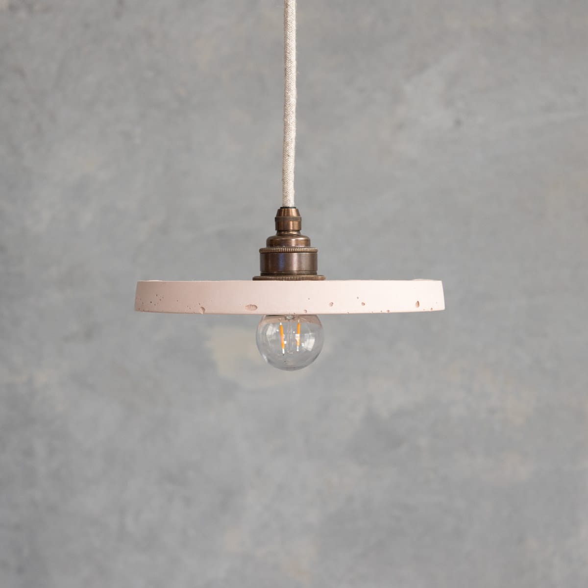 Loman Pendant in Nude. Image copyright of Crete Studio. Behind the scenes style image of woman holding a lighting pendant in front of a concrete wall while a man takes a picture on a phone.