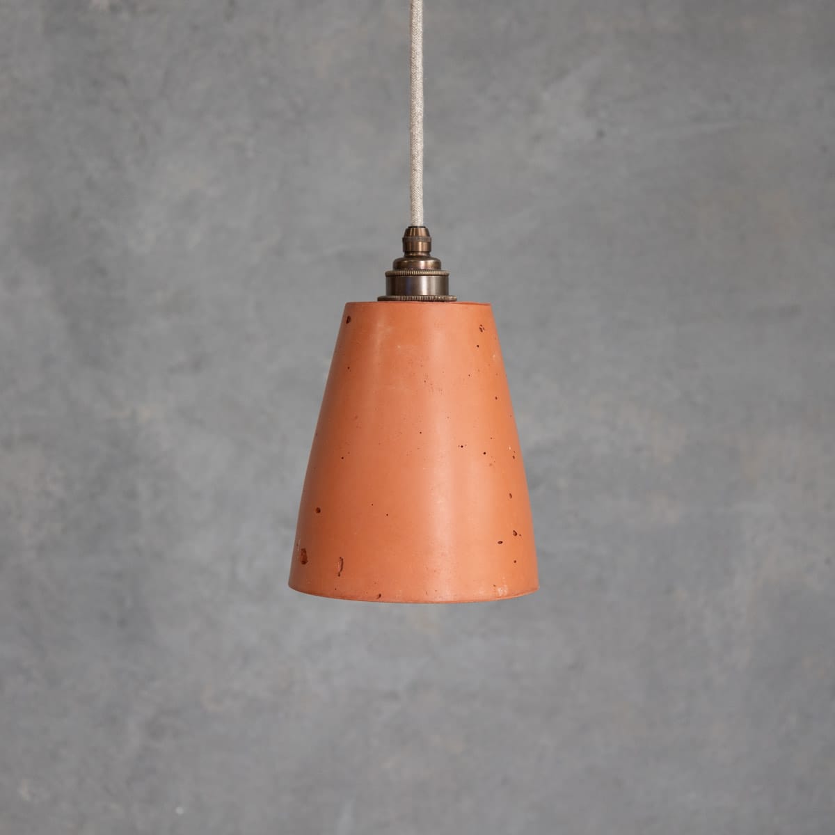 Maca Pendant in Terracotta. Image copyright of Crete Studio. Terracotta cylindrical pendant light hanging in front of concrete wall.