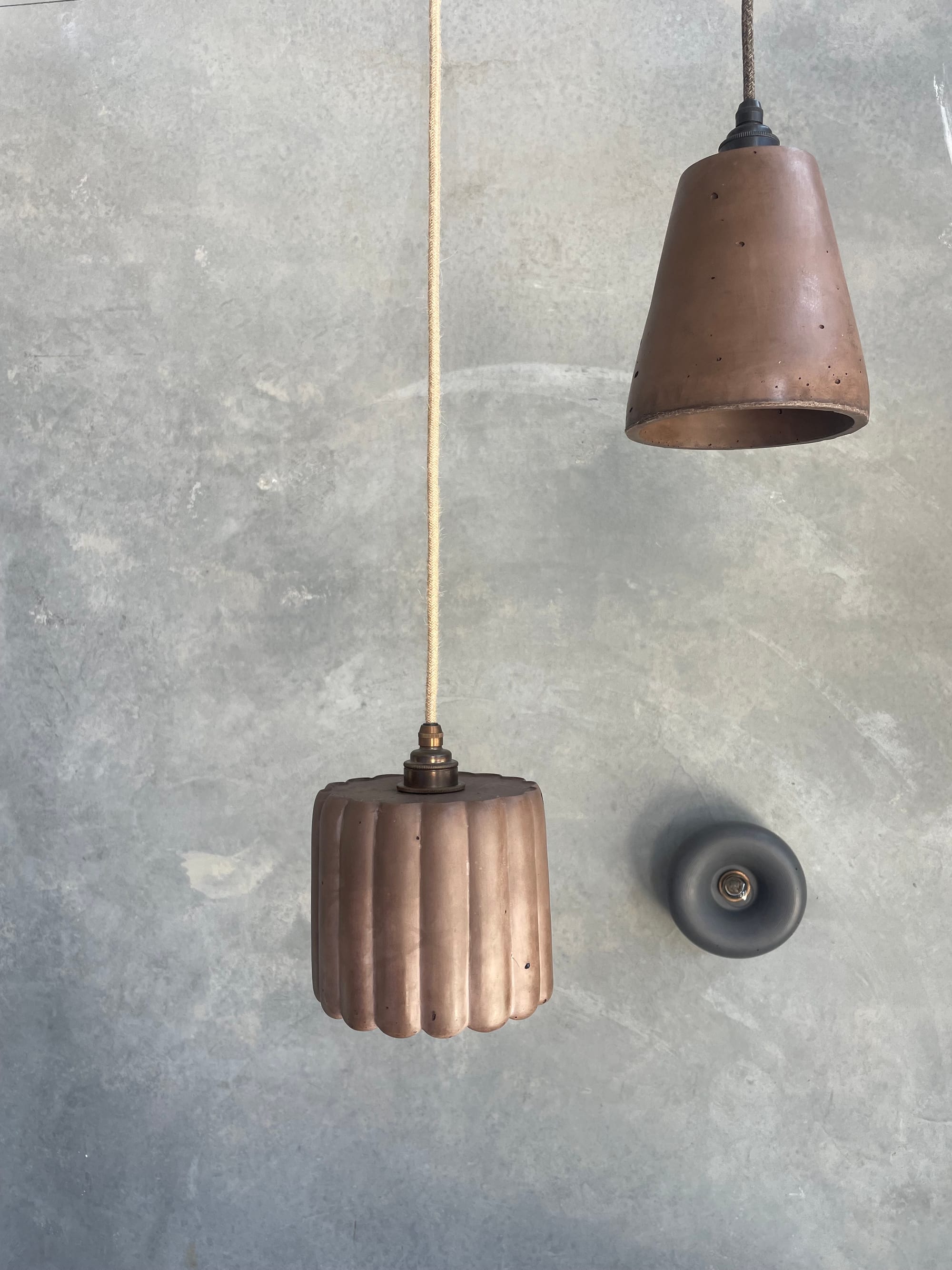 Crete Studio. Left to right: Large Fluted Pendant in Cocoa, Maca Pendant in Cocoa and Haze Wall Sconce in Charcoal. Two brown concrete pendants hanging on front of concrete wall. Small round grey wall fixture light on right.
