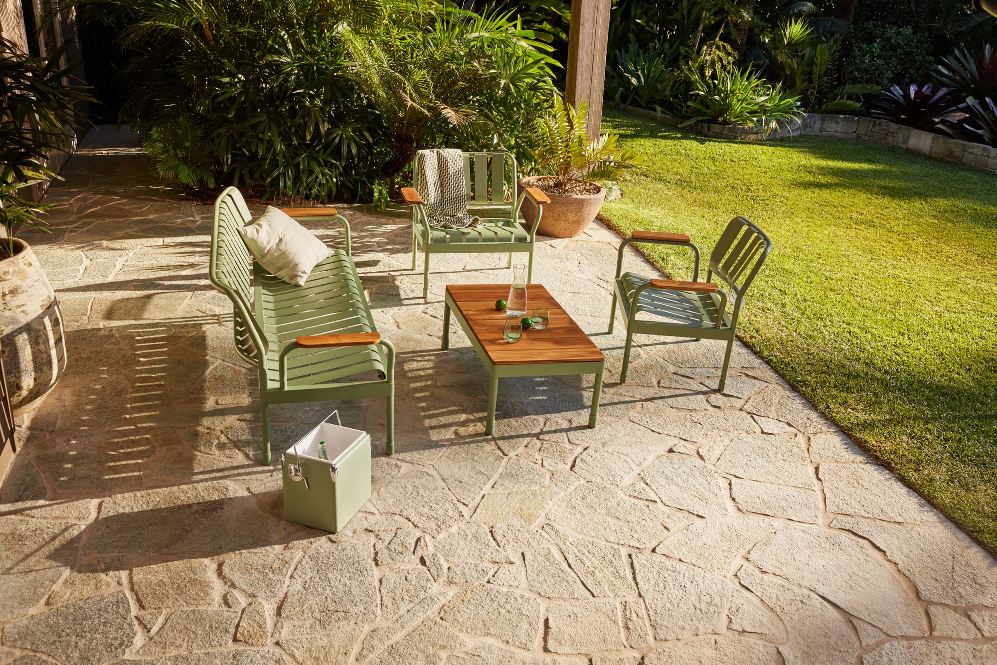 Torakina Outdoor Lounge Set by Koala in Soft Moss. Outdoor metal lounge chairs and dining chairs in green with metal and timber coffee table on paved floor with grass.