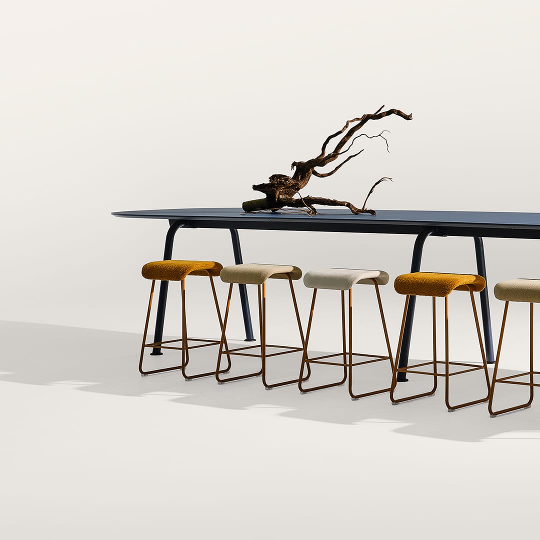 Sela Stool by EFG. Blue bench table in white space with five various coloured stools, with brass legs and white, tan and orange cushions. 