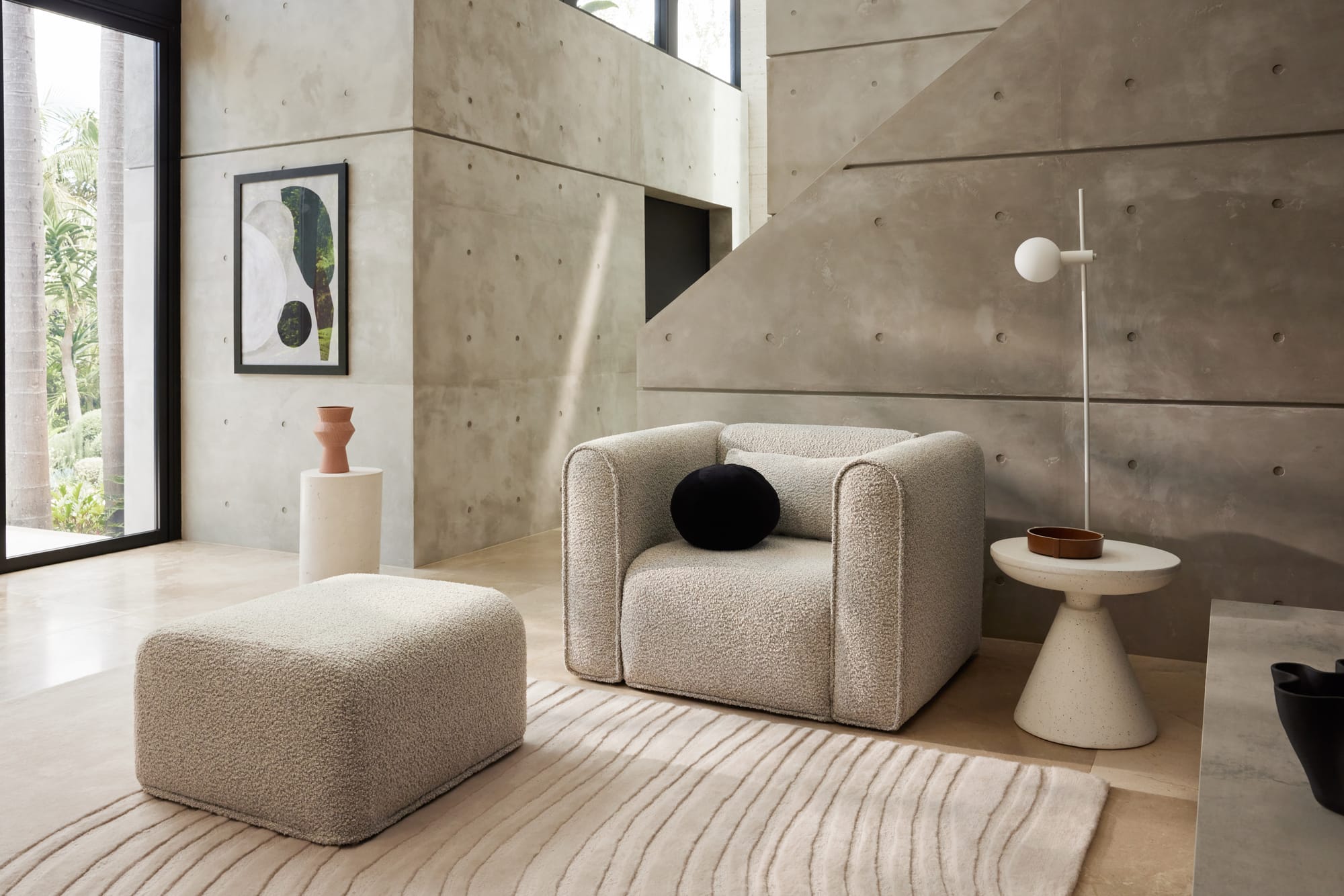 Bangalow Modular Sofa in Limestone. Cream boucle armchair in front of concrete panelled wall and staircase, with matching ottoman. 