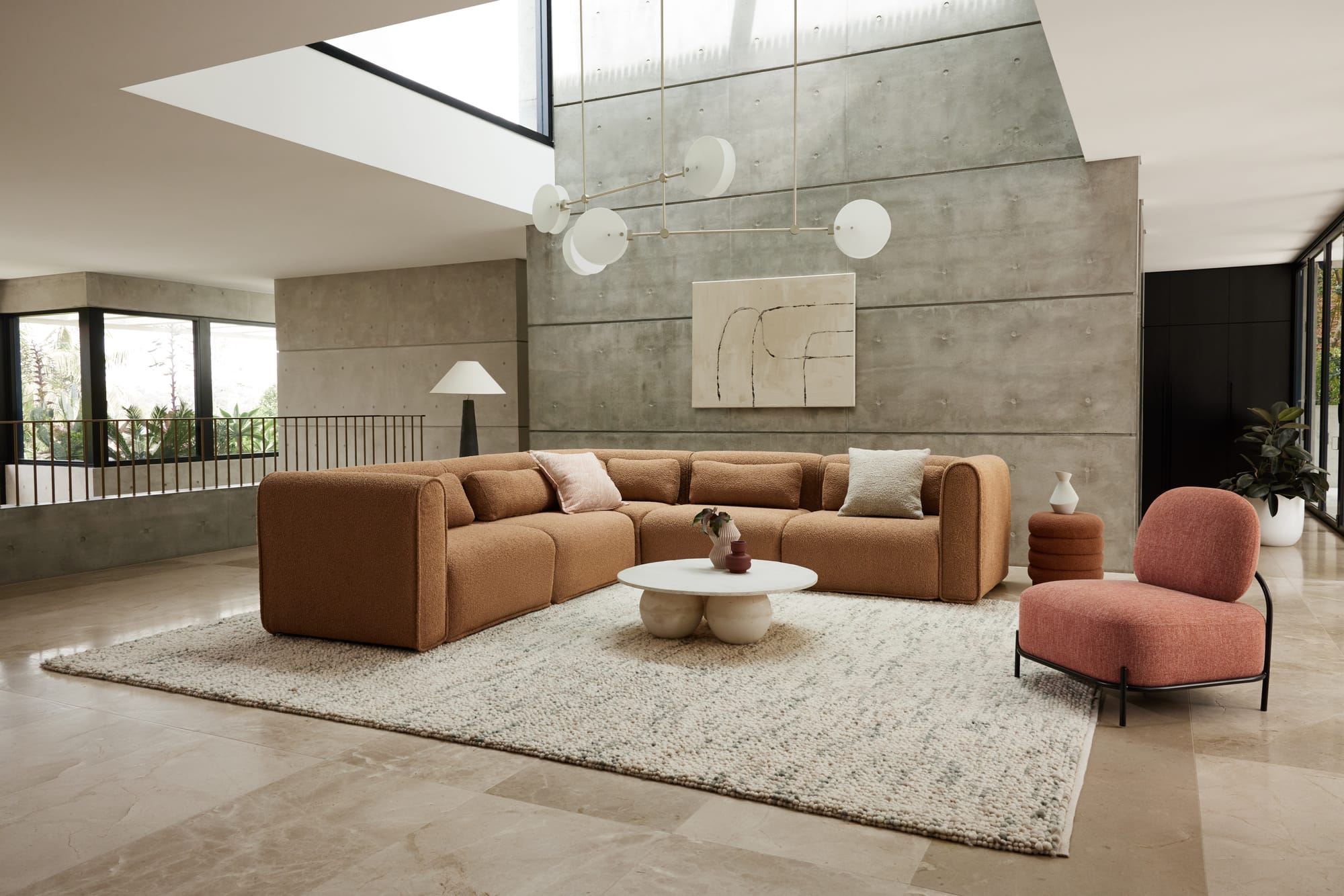 Bangalow Modular Sofa in Sand Dune. Tan boucle couch in L-arrangement in front of double heigh concrete paneled wall, with red boucle armchair and white coffee table in foreground. 