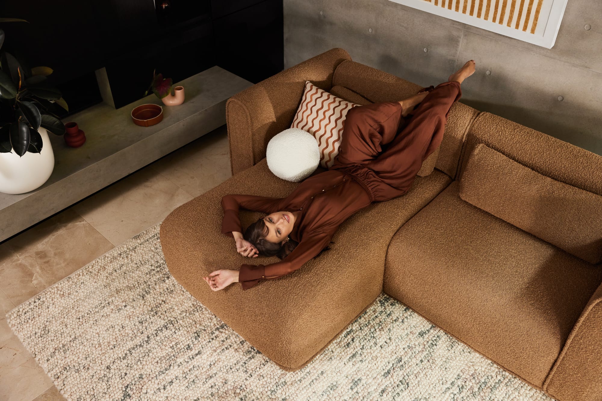 Bangalow Modular Sofa in Sand Dune. Woman in burgundy outfit laying on tan boucle sofa on knotted rug. 