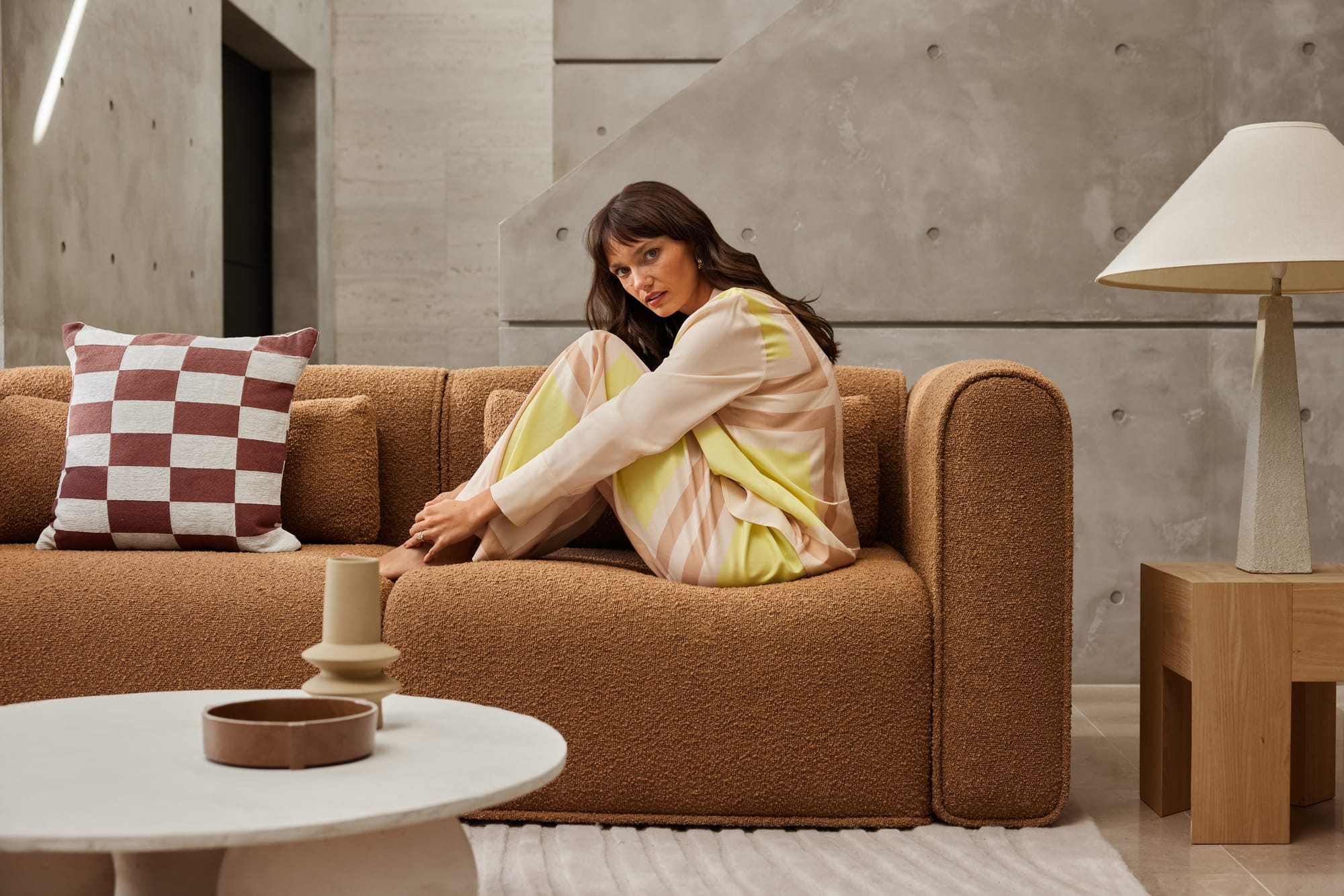 Bangalow Modular Sofa in Sand Dune.  Woman sitting on burnt orange boucle sofa with timber side table, white round coffee table in foreground and checkered burgundy and white cushion to the left. 