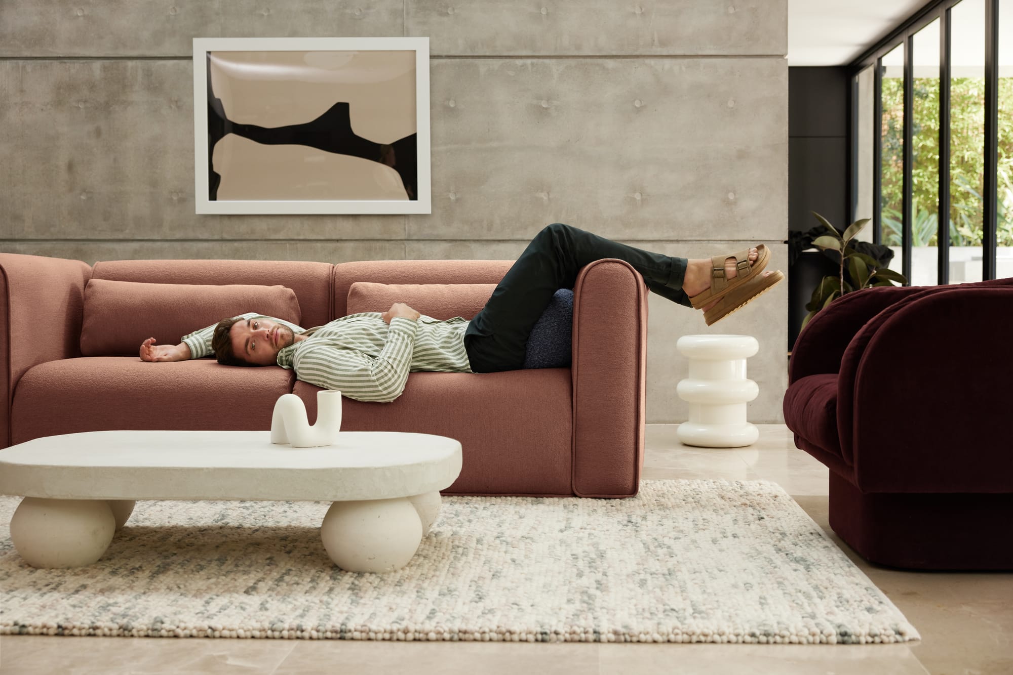 Bangalow Modular Sofa in Sunset Rust. Man in jeans and striped shirt laying on rust coloured boucle sofa with his feet hanging over the edge, in front of concrete wall and abstract white coffee table. 