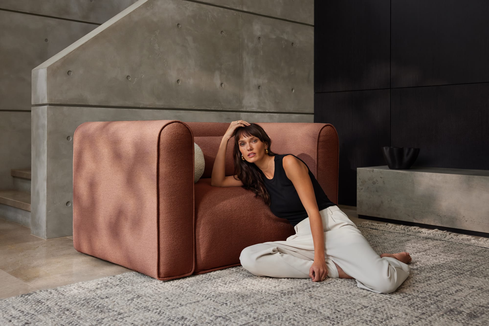 Bangalow Modular Sofa in Sunset Rust. Woman siting on knotted floor rug, leaning against rust coloured boucle armchair in front of concrete panel wall. 