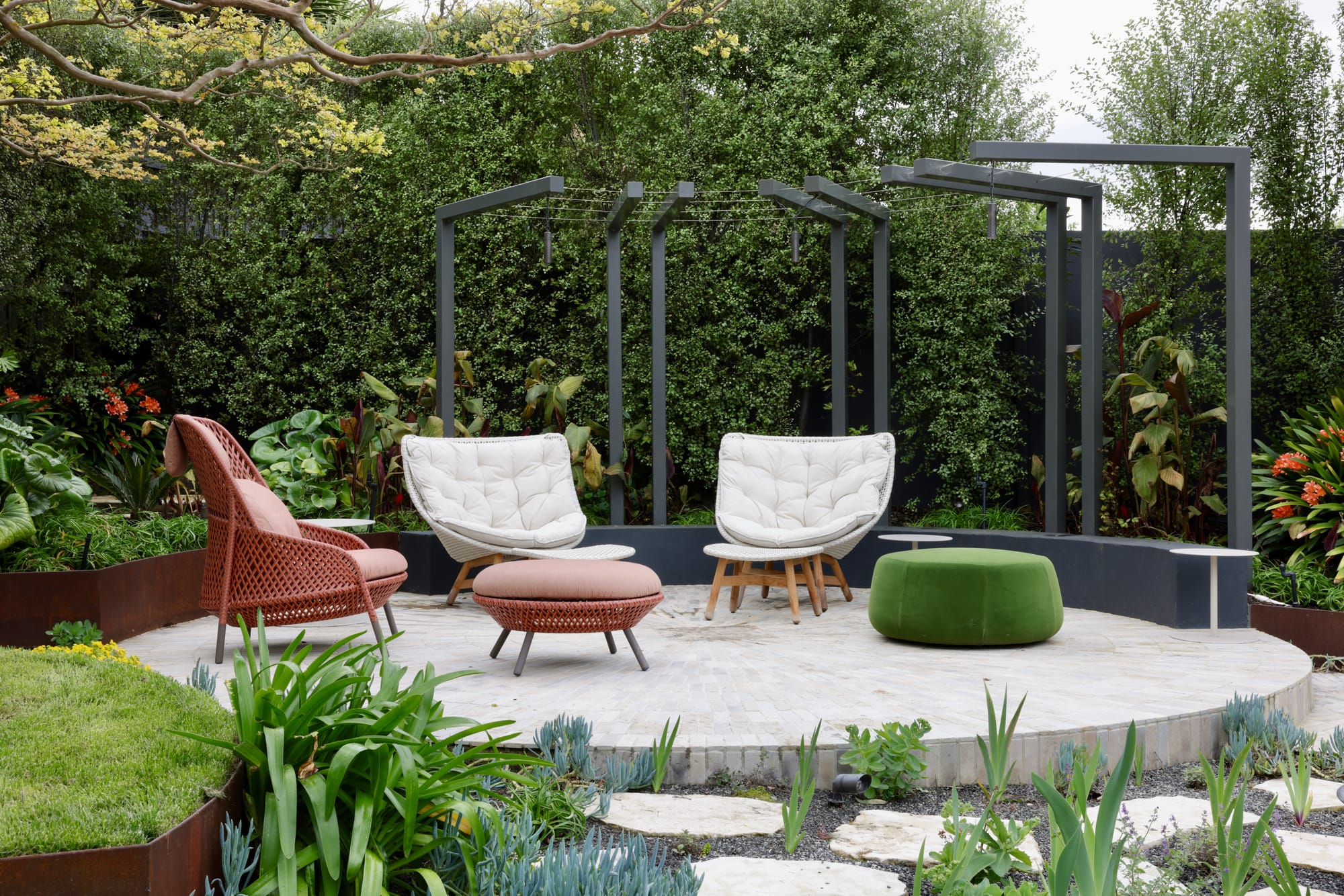 Glen Iris by Julie Crowe Landscape Design x Cosh Living. Photography by&nbsp;Eloise Sim. Two white armchairs and one red armchair with matching footstool arranged on white round platform in outdoor garden. Green ottoman and dark timber arbor to the right of platform with tall hedges on the left. 