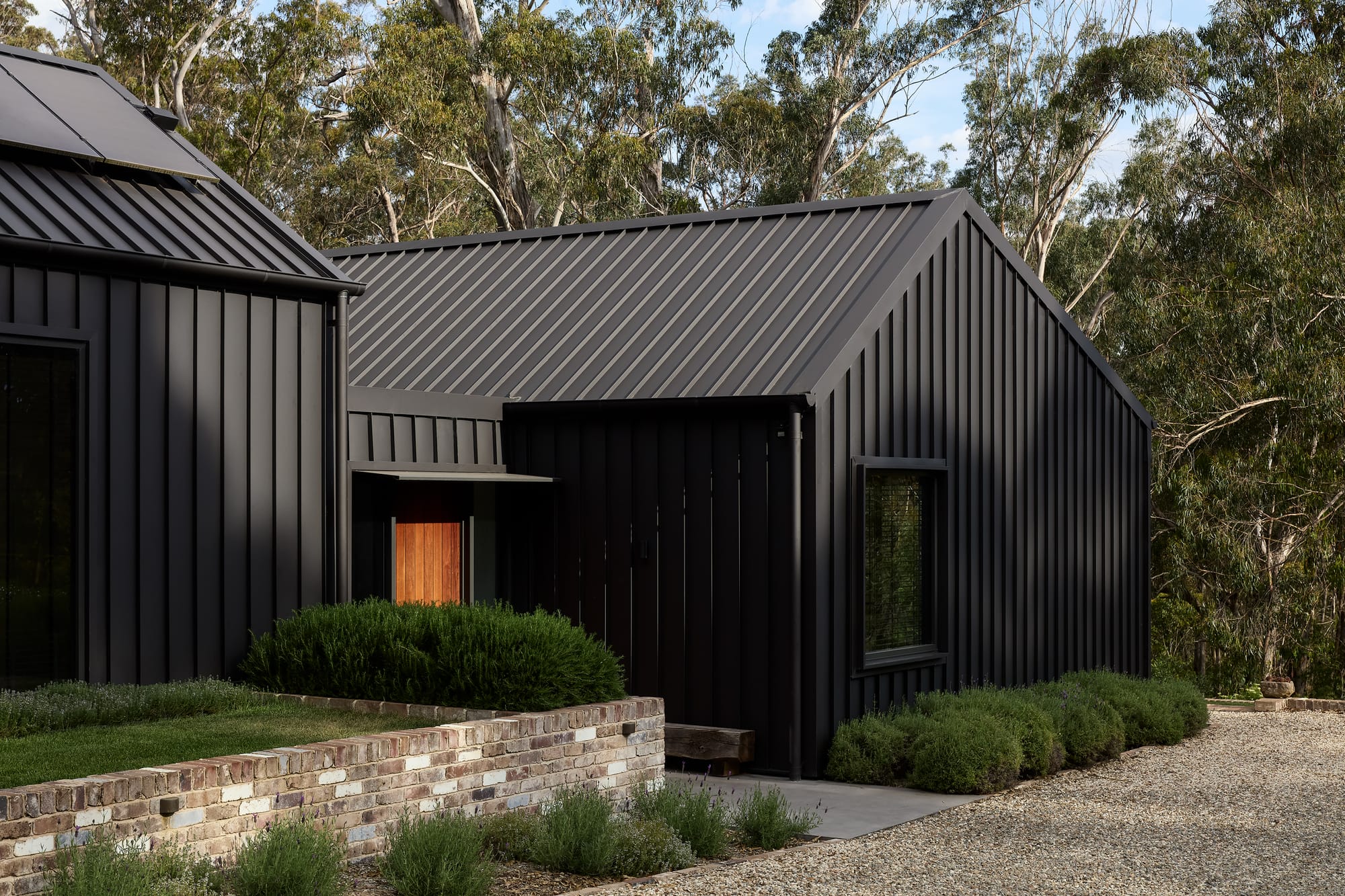 House Kimberley by AO Design Studio. Photography by Luc Remond. Black clad pavilion home with pitched roof, recycled brick garden beds with lush grass and shrubs, and crushed gravel driveway in front of native Australian bushland. 