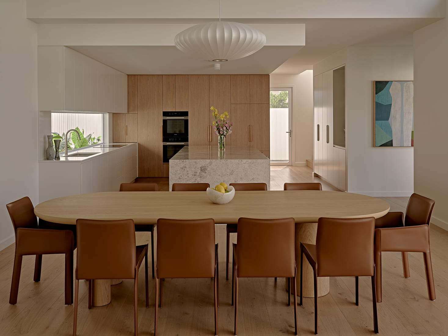 Twin Palms House by FURNISHD. Photography by Brock Beazley. Dining and kitchen space with long timber dining table, leather chairs, timber cabinetry and flooring, stone island bench and white pendant light. 
