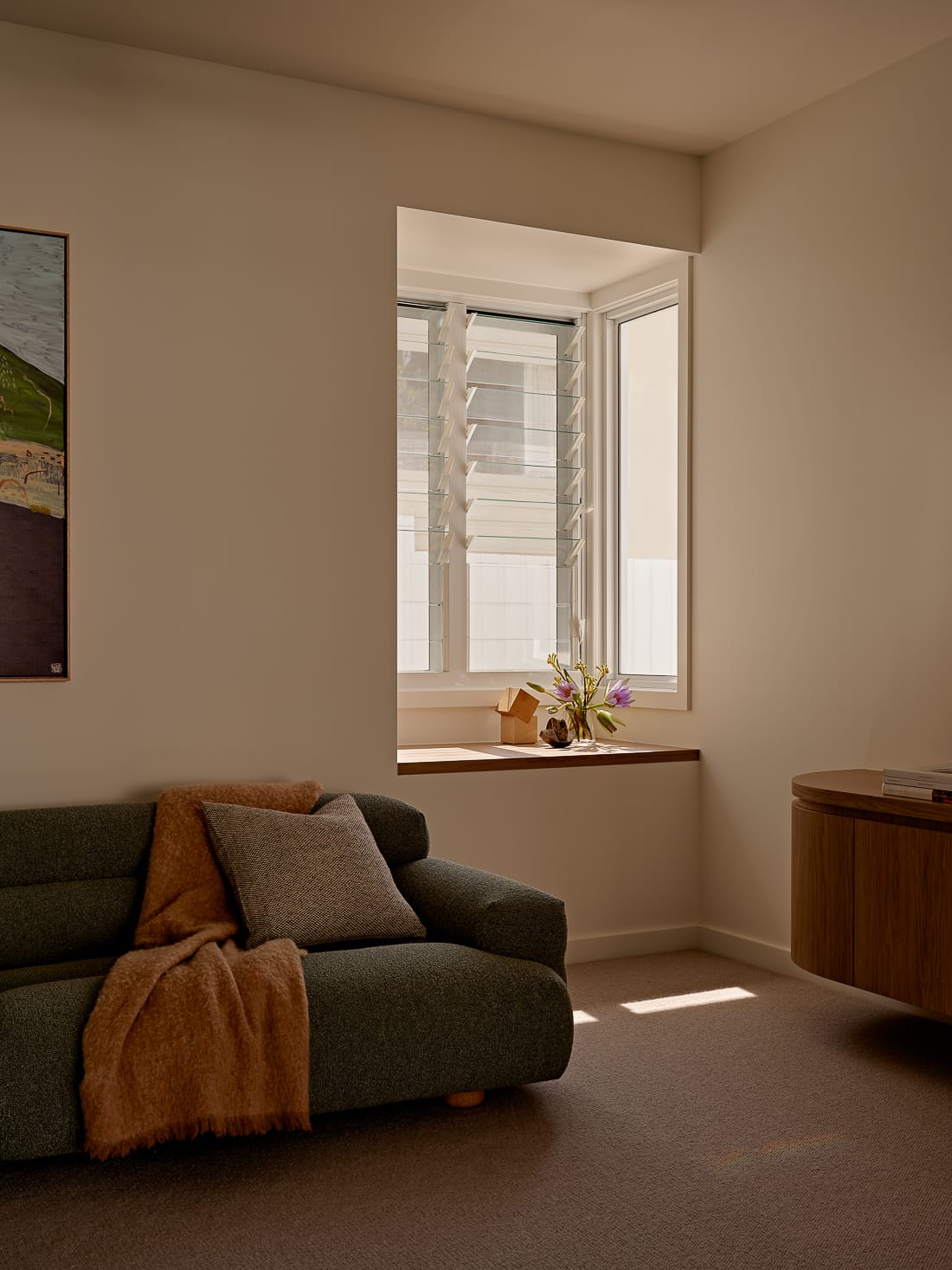 Twin Palms House by FURNISHD. Photography by Brock Beazley. Living space with small window alcove, green upholstered couch with orange throw, beige carpets and timber sideboard. 