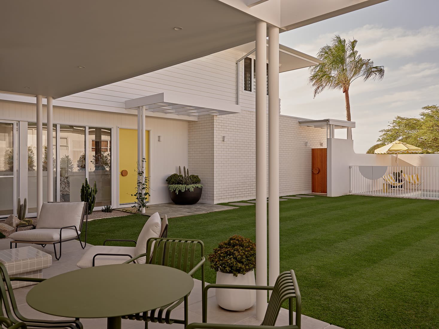Twin Palms House by FURNISHD. Photography by Brock Beazley. Backyard with undercover pergola with white and green metal furniture, green grass, pool in background and tall white brick walls.