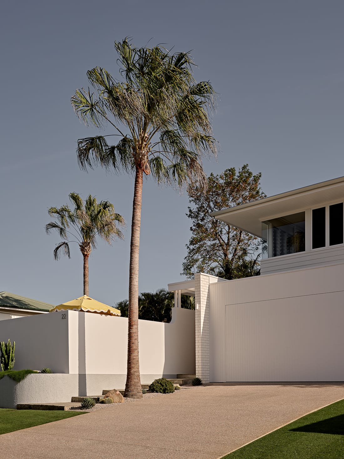Twin Palms House by FURNISHD. Photography by Brock Beazley. White double storey timber clad home with white concrete fence and paved driveway. Tall palm trees around property and yellow pool umbrella visible over fence.