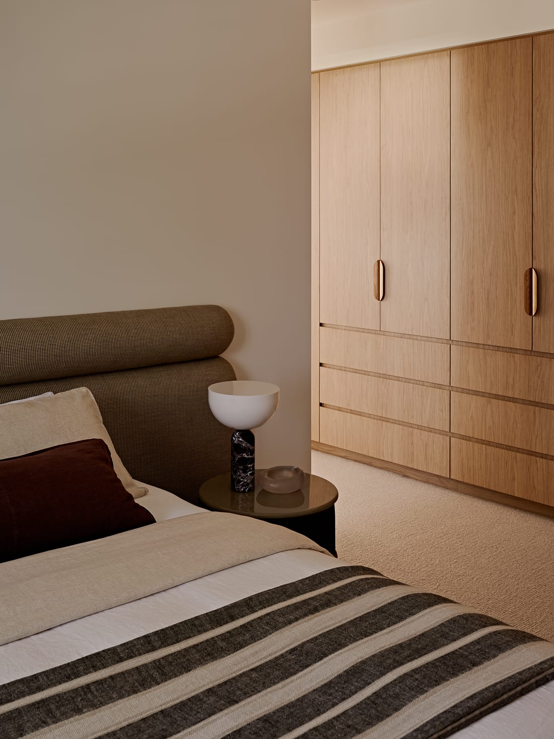Twin Palms House by FURNISHD. Photography by Brock Beazley. Bed with striped throw rug, upholstered bedhead and maroon throw cushions. Timber cabinetry in background and beige carpets.
