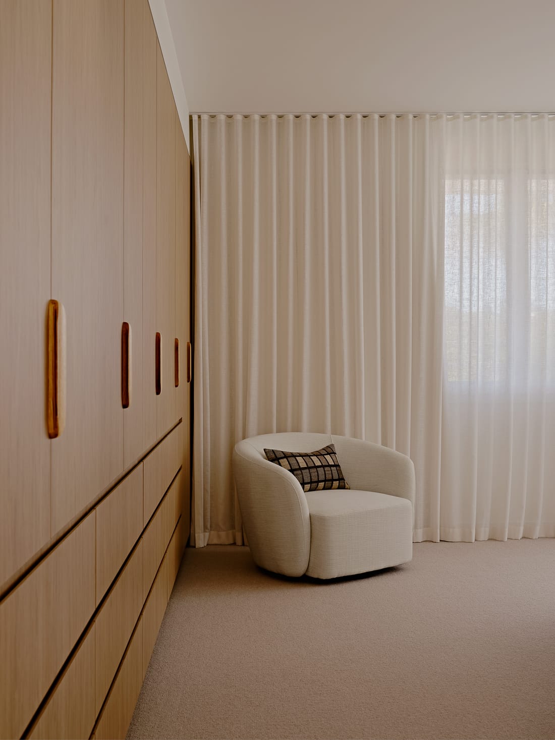 Twin Palms House by FURNISHD. Photography by Brock Beazley. White armchair in front of sheer full length curtains and timber cabinetry running from floor to ceiling.