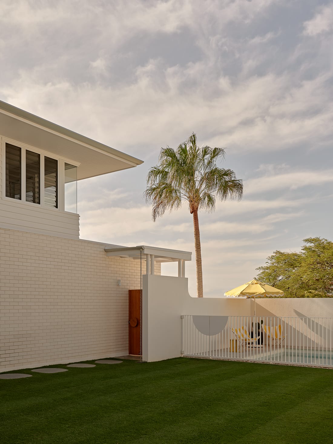 Twin Palms House by FURNISHD. Photography by Brock Beazley. Green lawn backyard with round step pavers, a white fence around a pool and a light brick wall. Dark red timber gate in fence. 