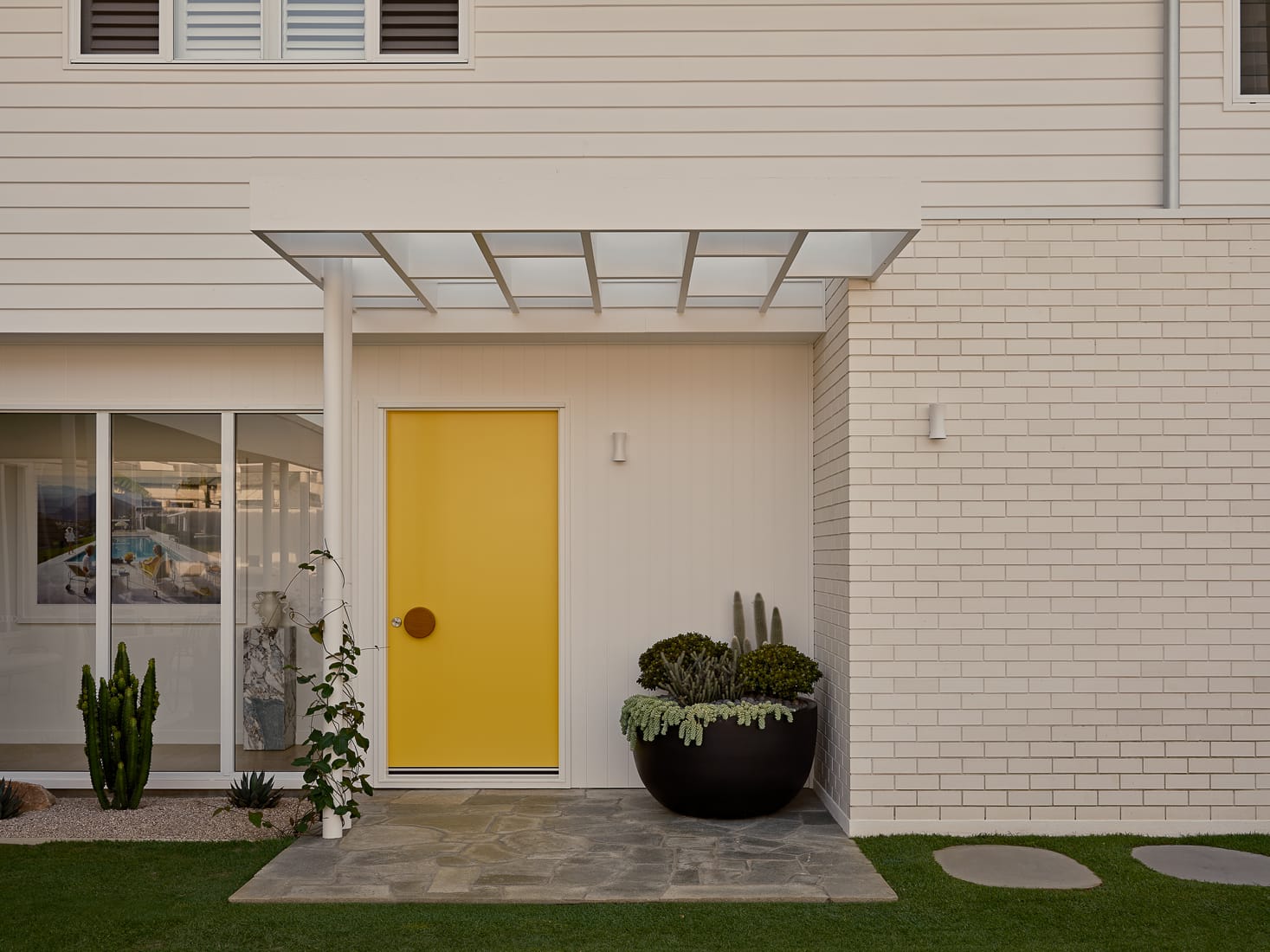 Twin Palms House by FURNISHD. Photography by Brock Beazley. Painted yellow front door with timber round knocker, pergola roof, paved floor, white brick walls and timber clad second storey.