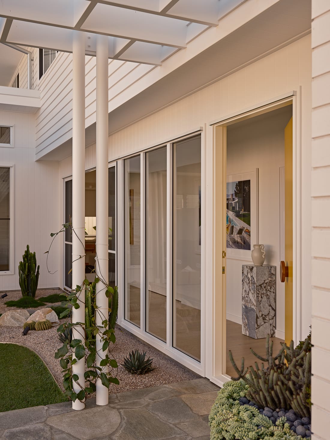 Twin Palms House by FURNISHD. Photography by Brock Beazley. Full length glass windows line hallway, with small courtyard garden on the left and exterior of the interior walkway. Two simple metal poles support pergola roof outside, with vines climbing up them.