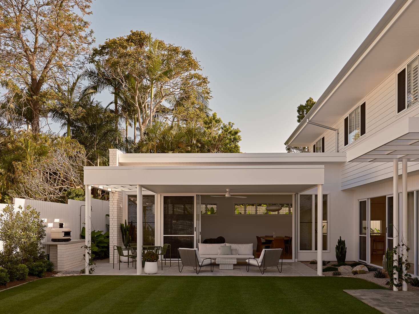 Twin Palms House by FURNISHD. Photography by Brock Beazley. Exterior patio with white walls and roof, green grass in foreground and plants scattered throughout.