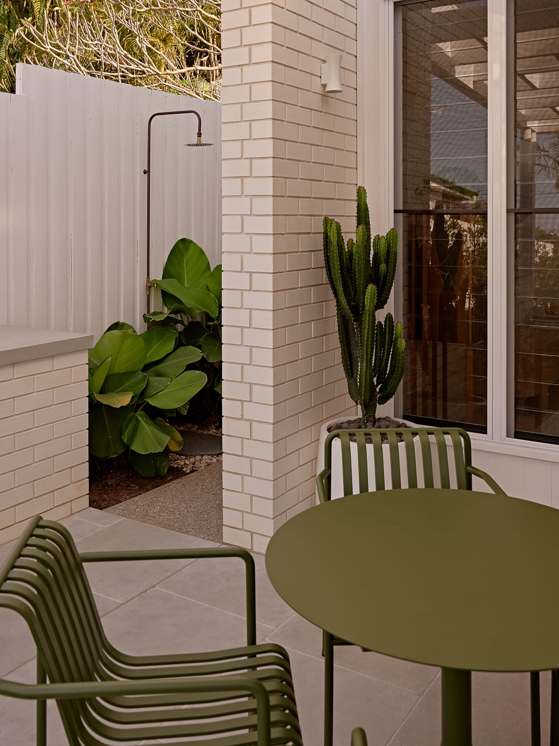 Twin Palms House by FURNISHD. Photography by Brock Beazley. Moss green metal outdoor furniture on grey paved patio, with white brick walls and garden in background. 