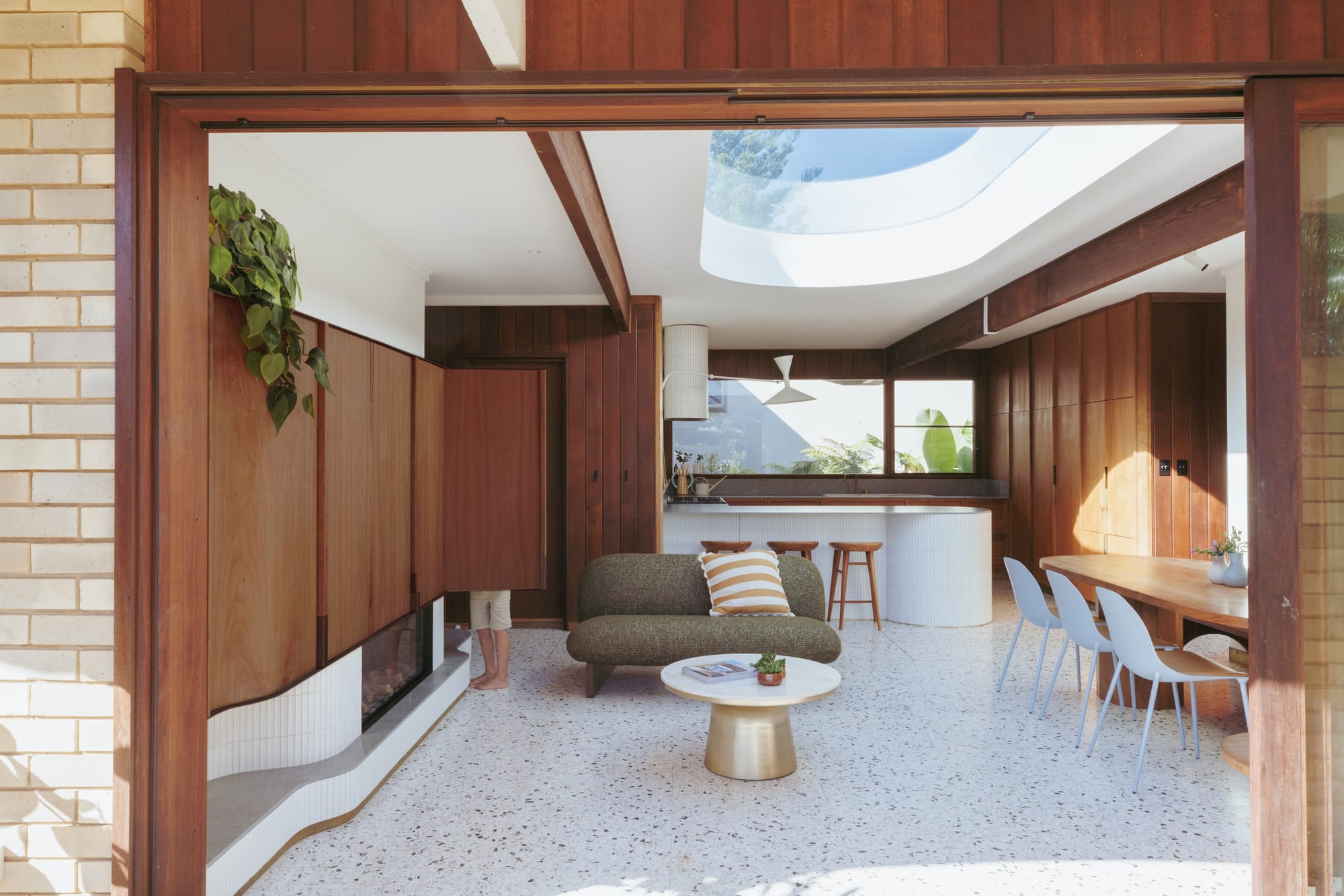 Casaballin by buck&simple. Photography by Tim Pascoe. Open plan living, kitchen and dining space with white terrazzo flooring, mid-century modern curved timber cabinetry, curved skylight and textured furnishings in chrome and olive colourways. 