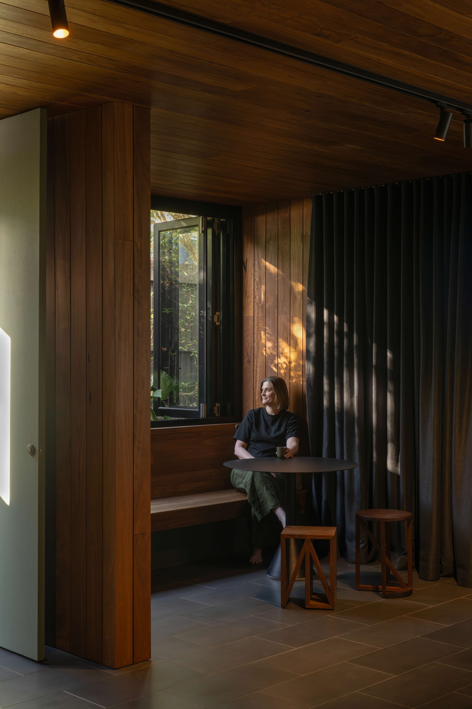 Cornerstone by Loupe Architecture. Photography by Alanna Jayne McTiernan. Integrated timber bench seating, with dark tile flooring, dark curtains, timber stools and large window overlooking the garden. 