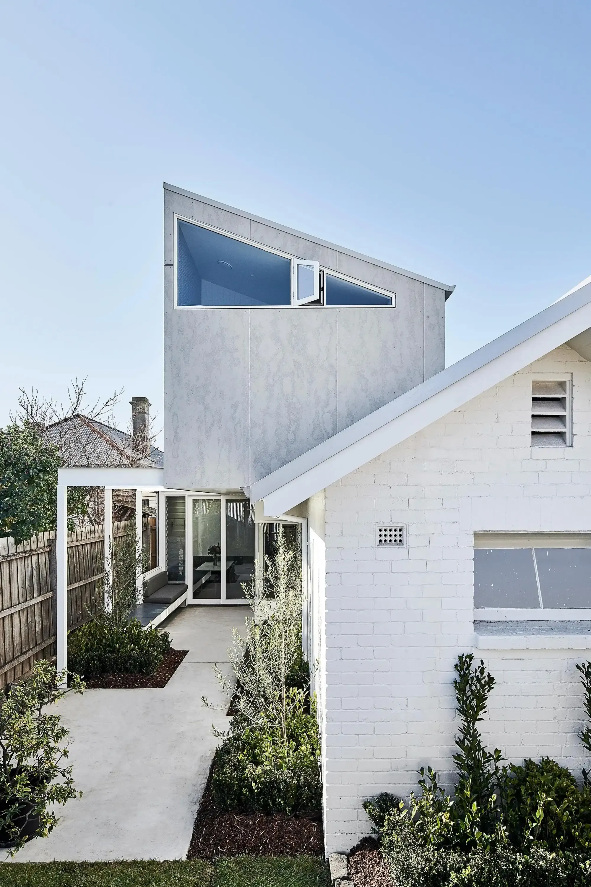 Dairy House by True Story. Photography by Dean Bradley Photography. Rear facade of multilevel residential home with white brick lower level and cement clad upper floor with slanting roofline and garden beds. 