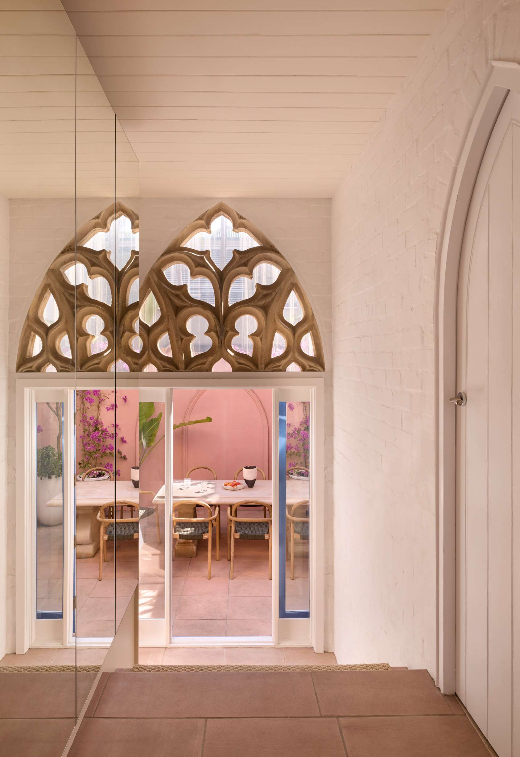 Chapel House by FURNISHD. Photography by Alicia Taylor. Hallway leading to arched doorway with cathedral features, opening onto paved courtyard with pink walls, stone table and timber dining chairs.