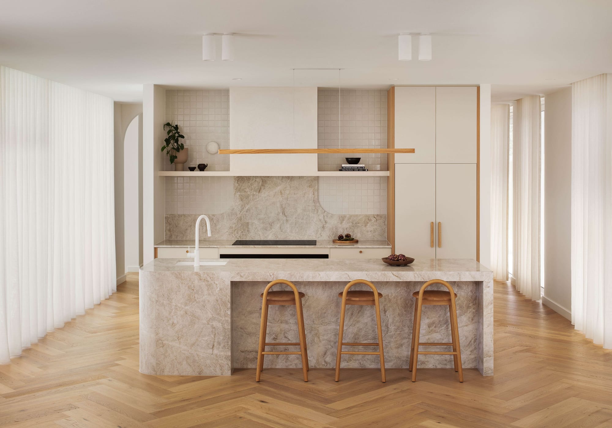 Haka House by Taouk Architects. Photography by Emily Bartlett. Kitchen with herringbone timber flooring, marbled stone benchtops and splashback, warm timber detailing and curved tiled accents. 