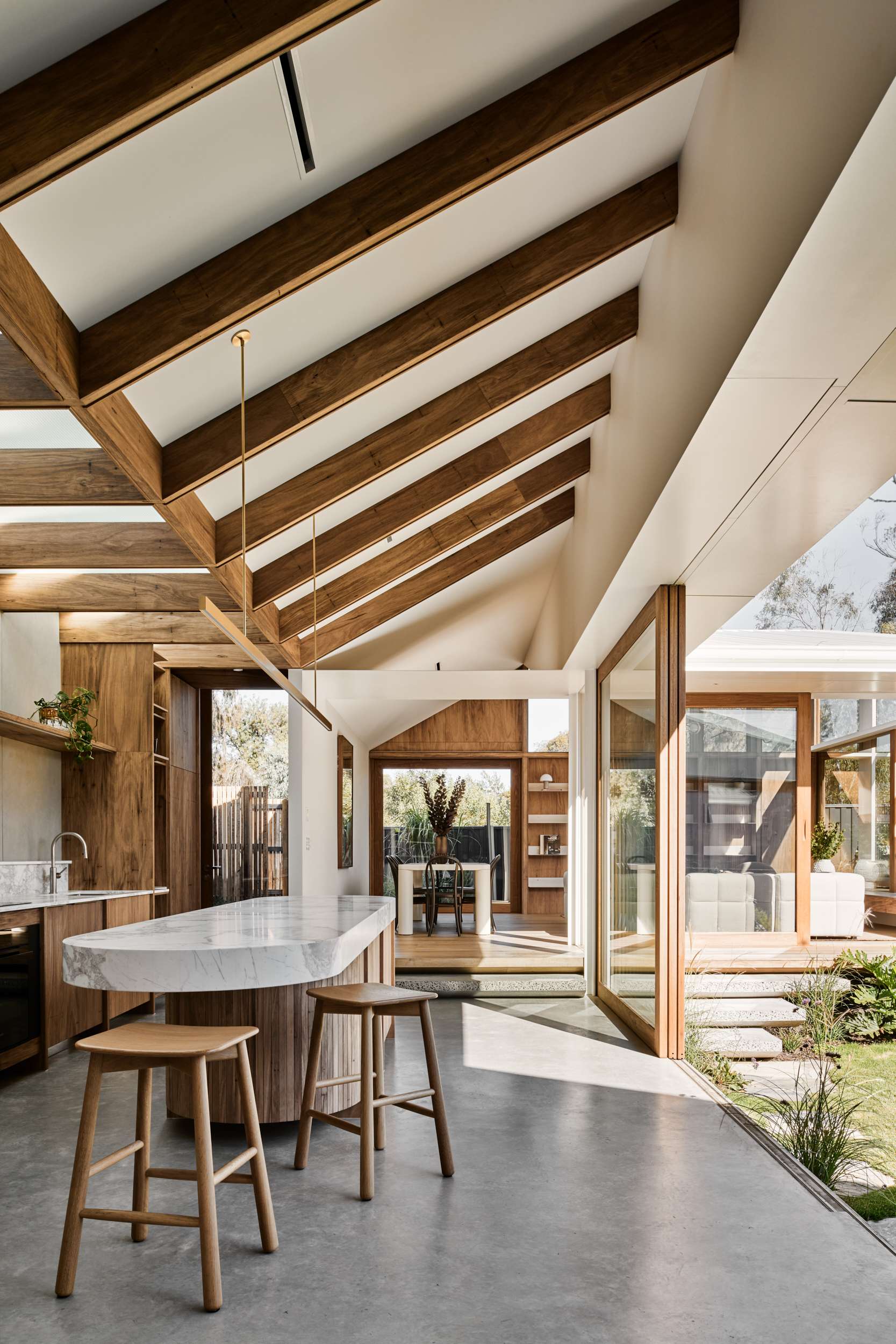 HUG HOUSE by FIGR Architecture Studio. Photography by Tom Blachford. Stepped open plan kitchen and dining space with polished concrete and timber flooring, raked ceiling with exposed beams, and full height timber sliding doors opening onto a grassed courtyard area. 