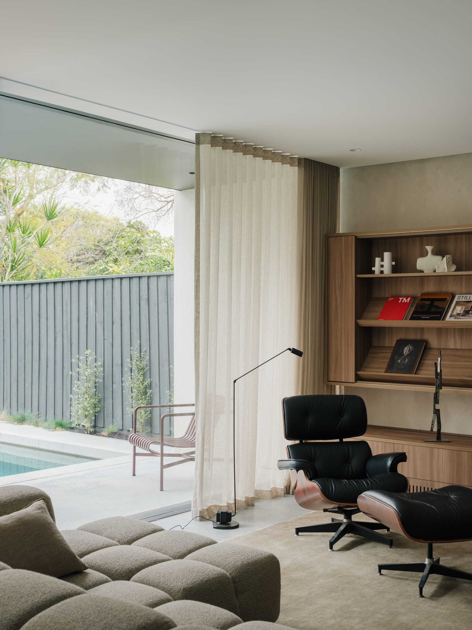 Petersham House by Cateaux Architects with CHROFI. Photography by Clinton Weaver. Living space with full lenth windows overlooking patio and pool, black Eames lounge chair and integrated open timber wall storage. 