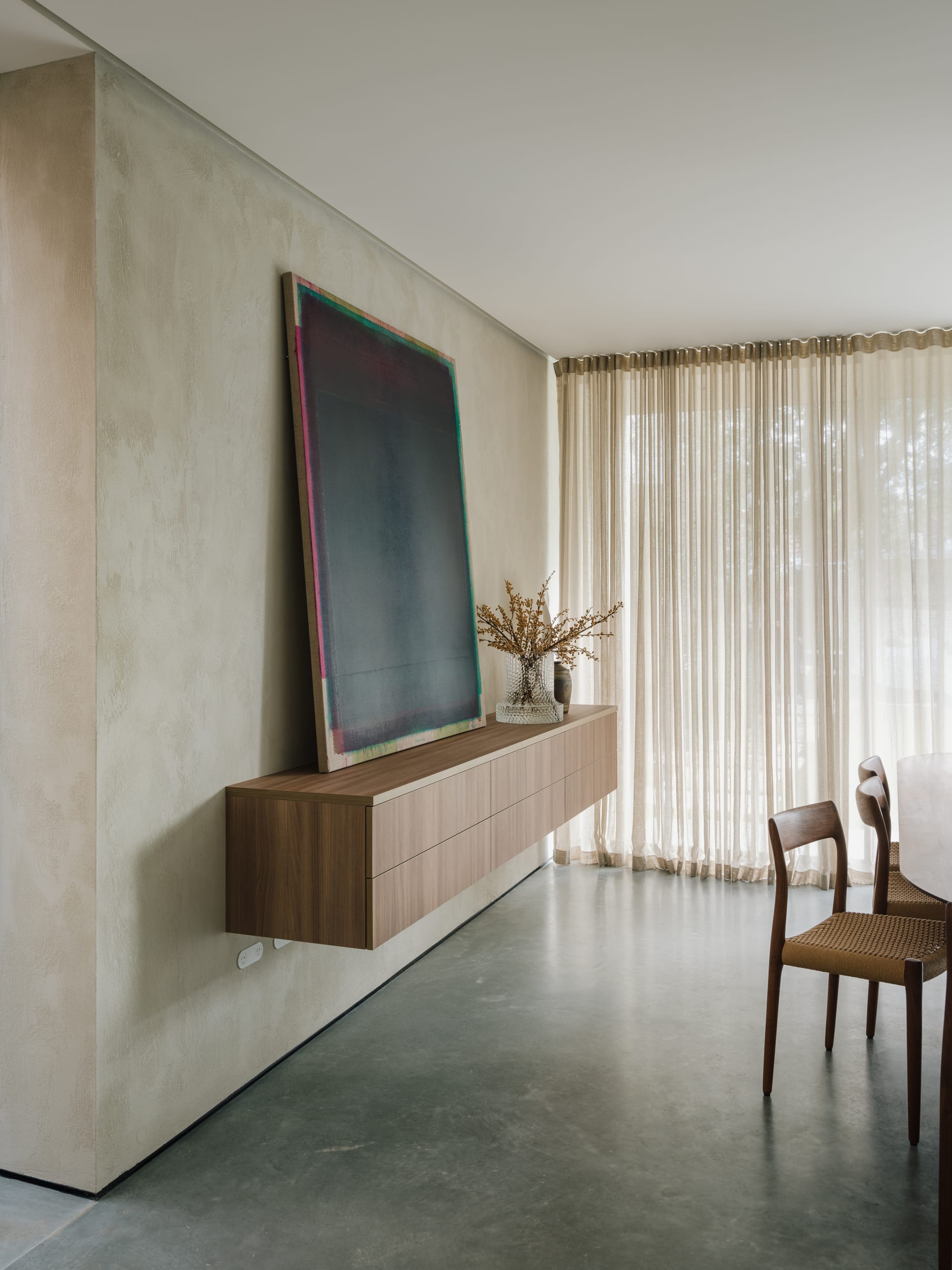 Petersham House by Cateaux Architects with CHROFI. Photography by Clinton Weaver. Floating timber sideboard with large abstract artwork in minimal room with polished concrete floors, limewashed walls, rattan chairs and sheer curtains. 