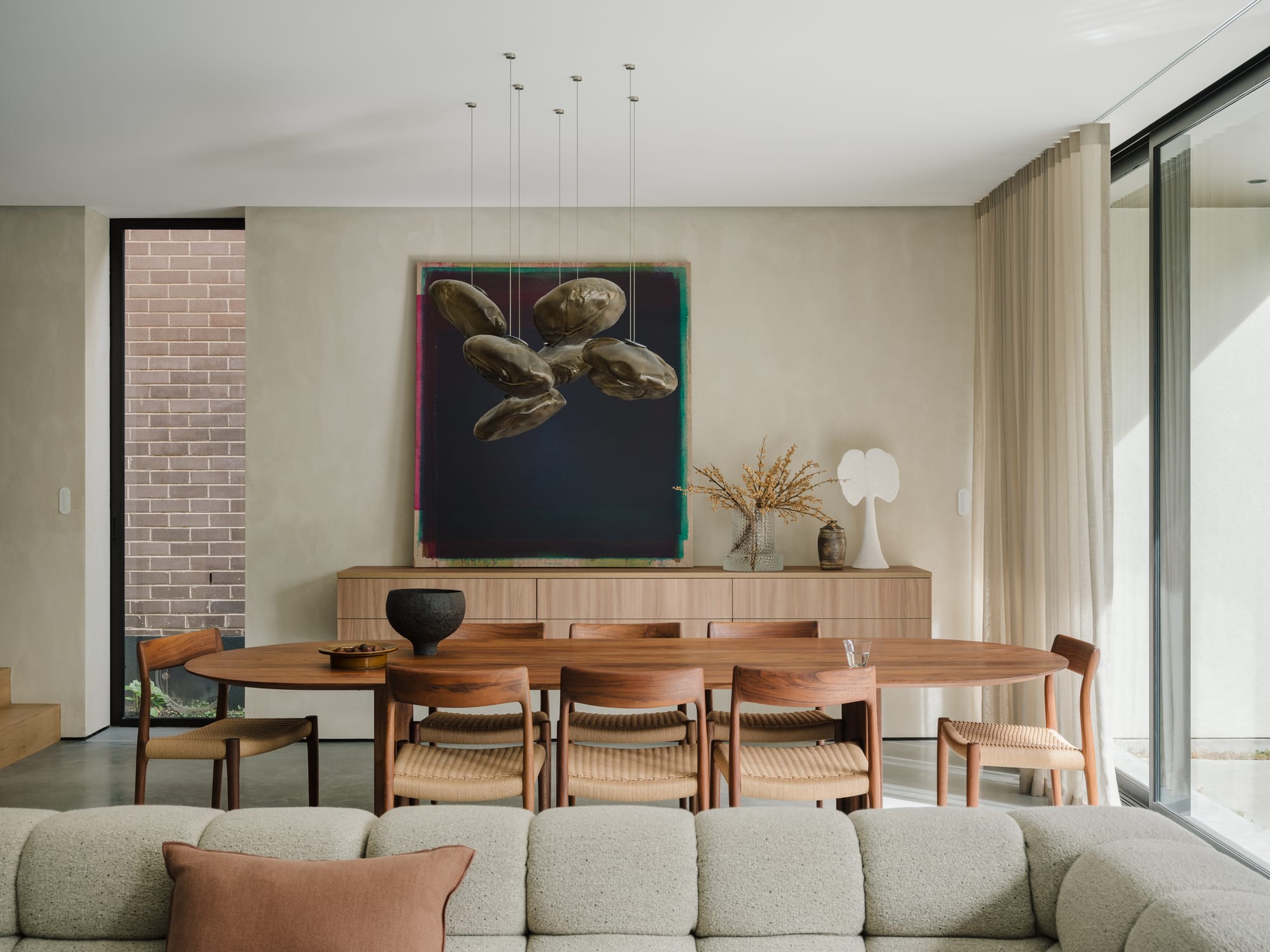 Petersham House by Cateaux Architects with CHROFI. Photography by Clinton Weaver. Oval timber dining table with rattan dining chairs in open-plan space with limewashed walls, grey tufted lounge, polished concrete floors, abstract pendants and sheer curtains. 