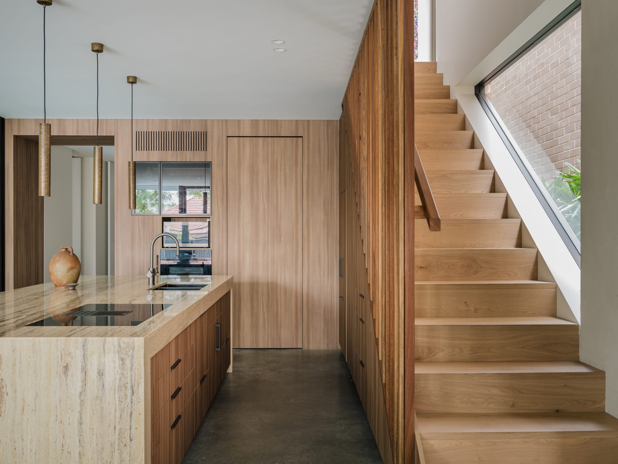 Petersham House by Cateaux Architects with CHROFI. Photography by Clinton Weaver. Kitchen with timber cabinetry, polished concrete flooring, stone island bench and timber staircase to the right with large window. 
